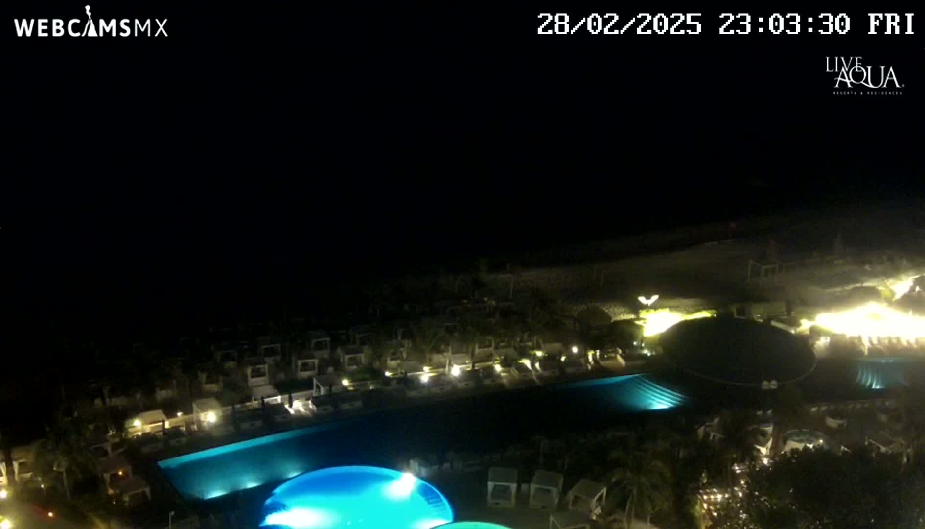 A nighttime aerial view shows a resort with illuminated pools and buildings near a beach under a dark sky.