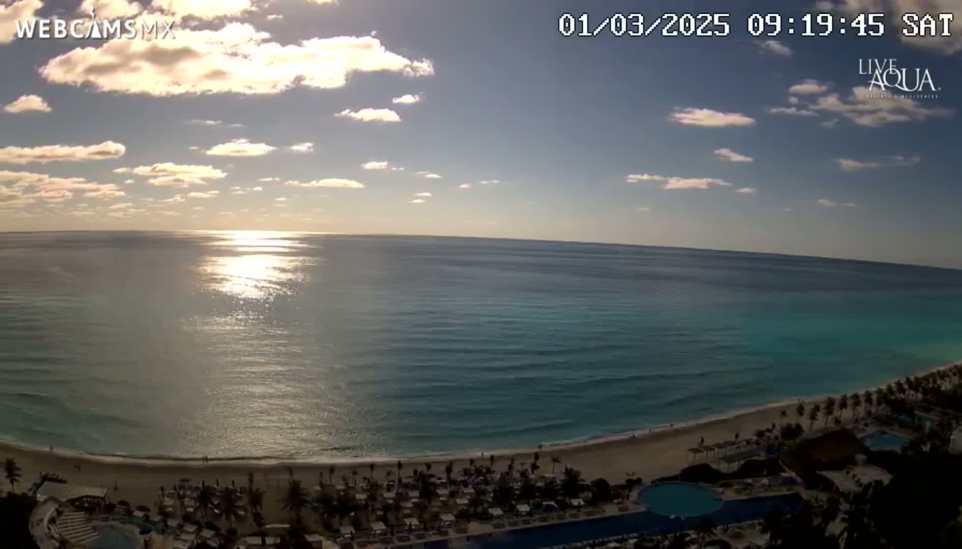 A mostly sunny day shows a beach with turquoise water and resort buildings under a partly cloudy sky.