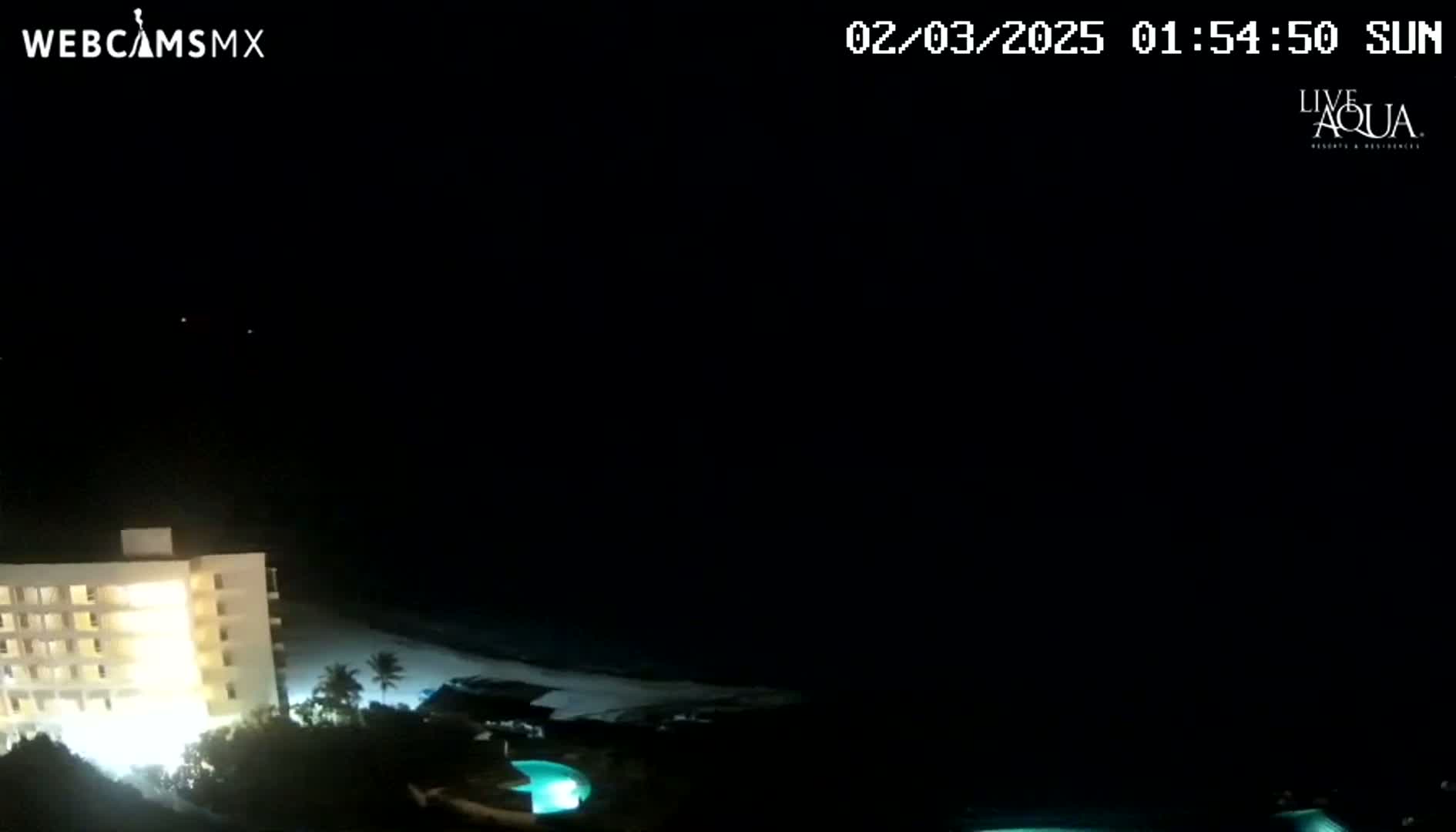 A brightly lit building sits near a beach at night, with a pool visible nearby under a clear, dark sky.