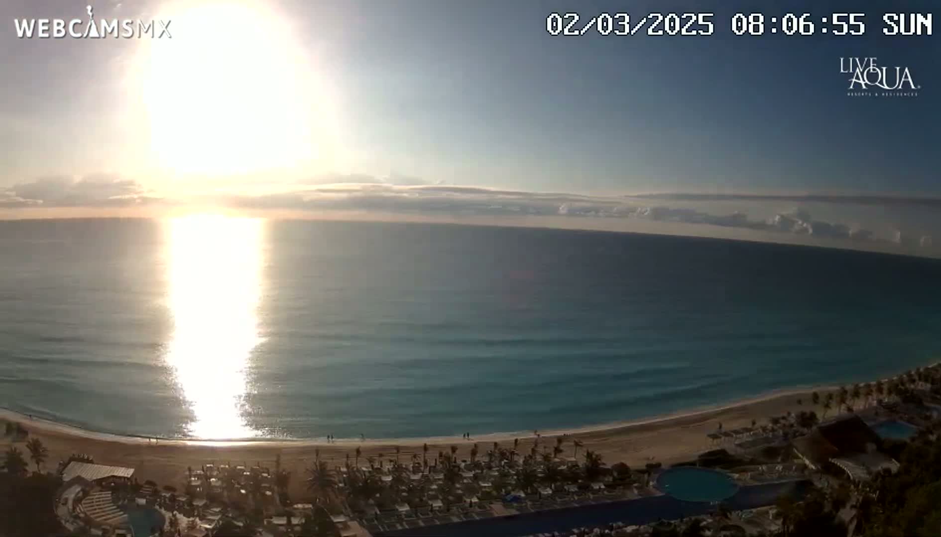 A bright sunrise over a calm turquoise ocean, shining onto a sandy beach with beach chairs and resort buildings.