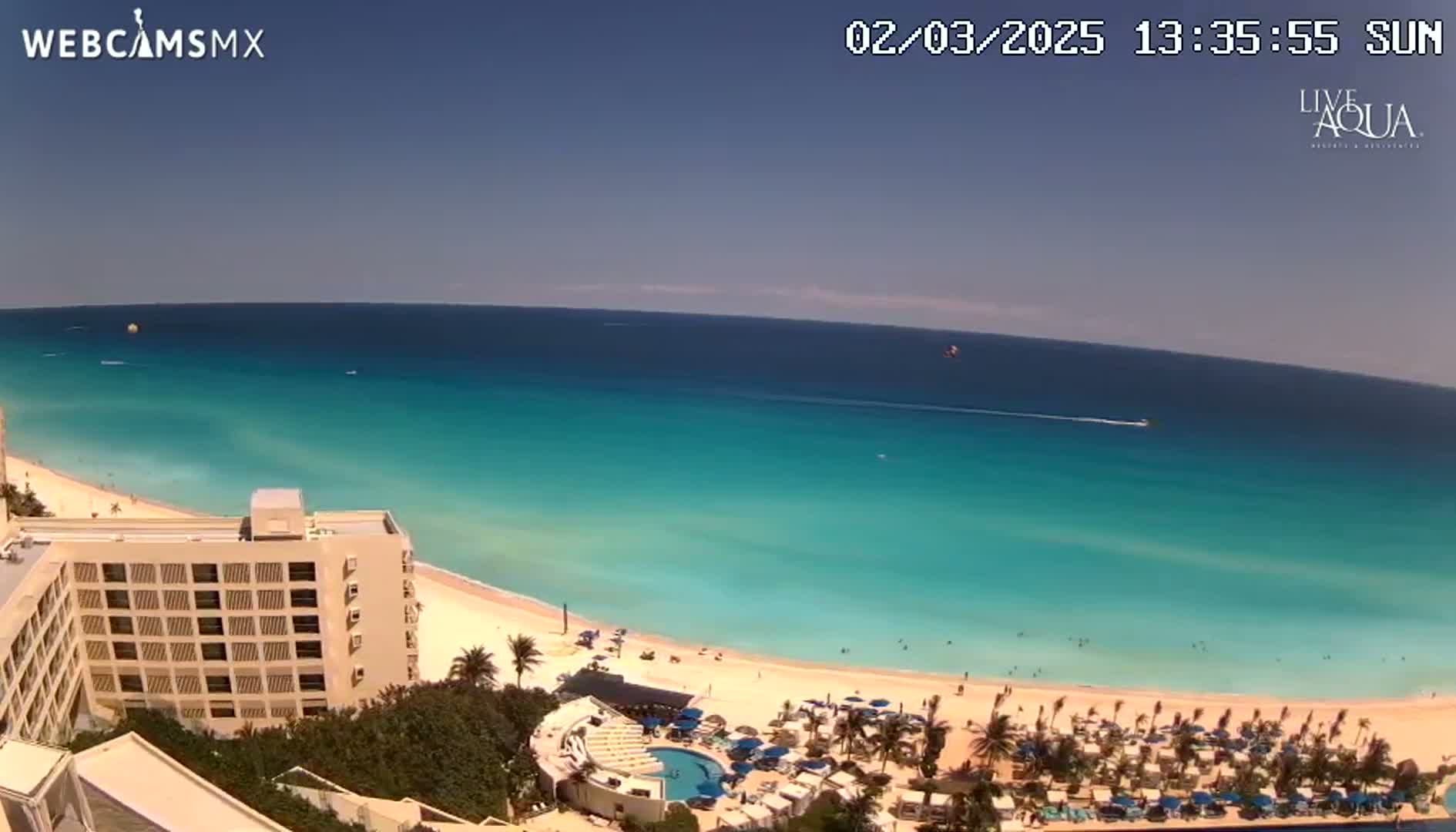 A high-angle view shows a beach resort on a sunny day with turquoise water and a white sandy beach, featuring a large building and several palm trees.