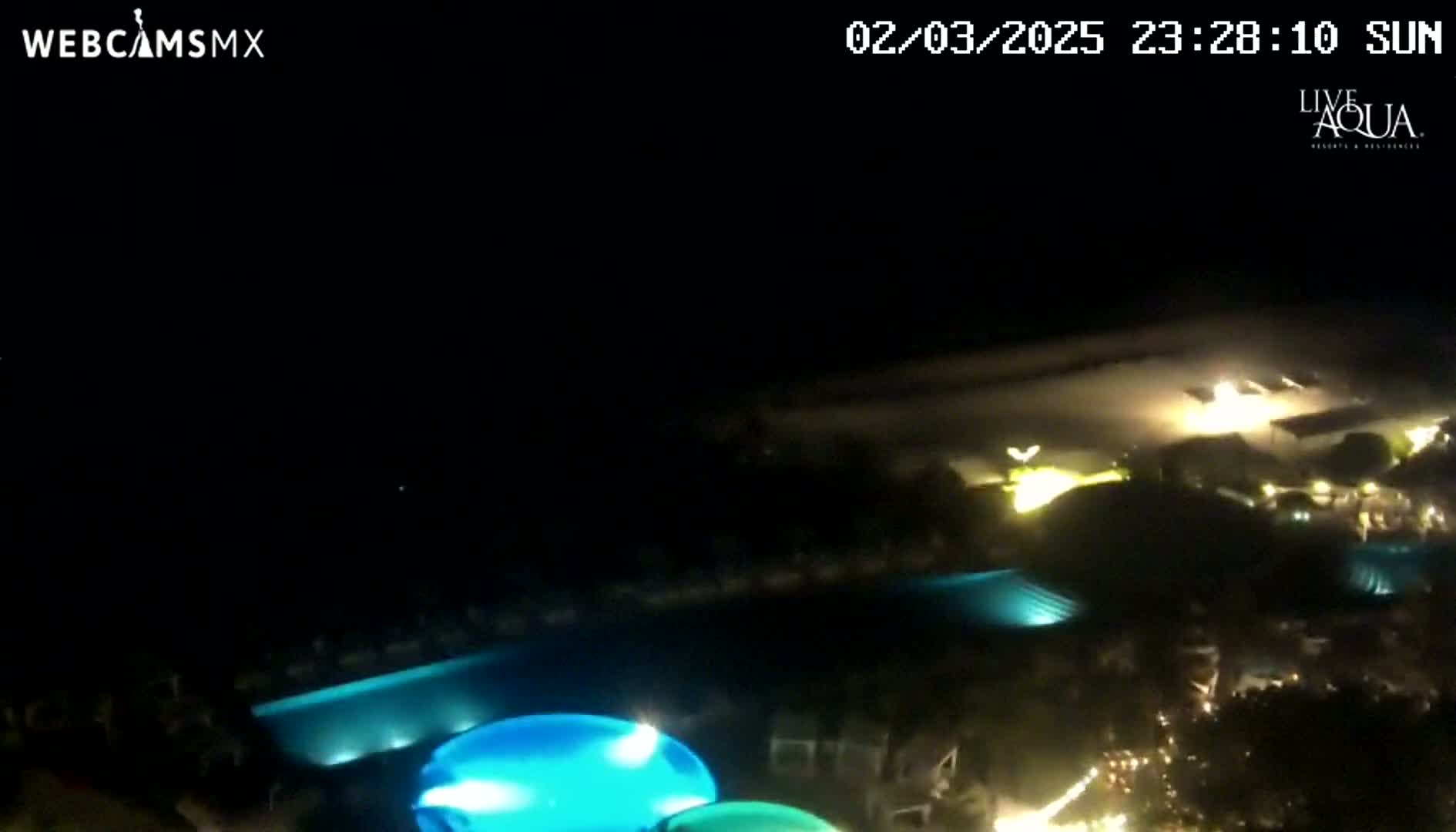 An aerial nighttime view shows a resort with illuminated swimming pools and surrounding landscaping under a clear, dark sky.