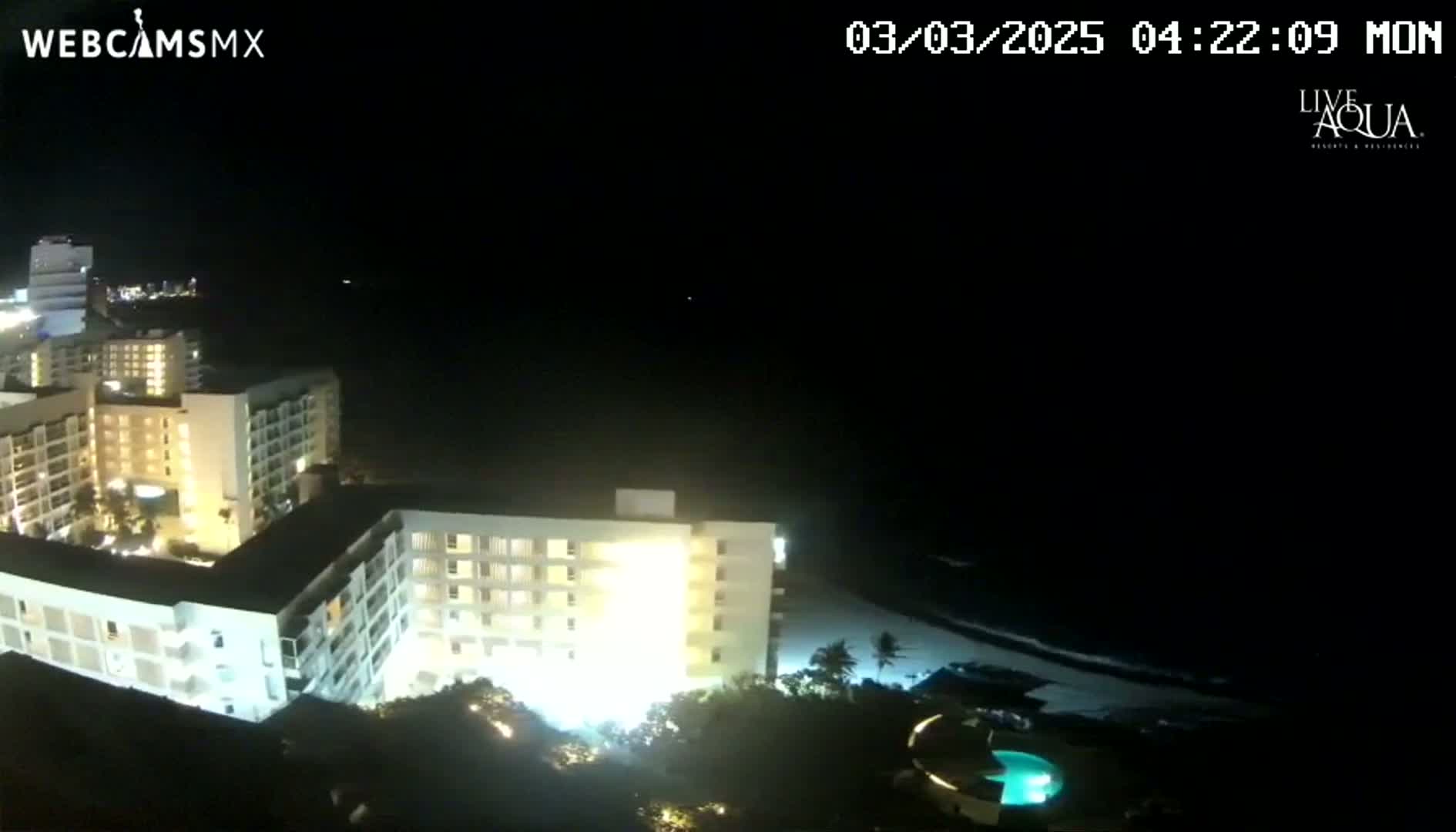 A nighttime aerial view shows brightly lit hotel buildings near a dark ocean under a clear sky.