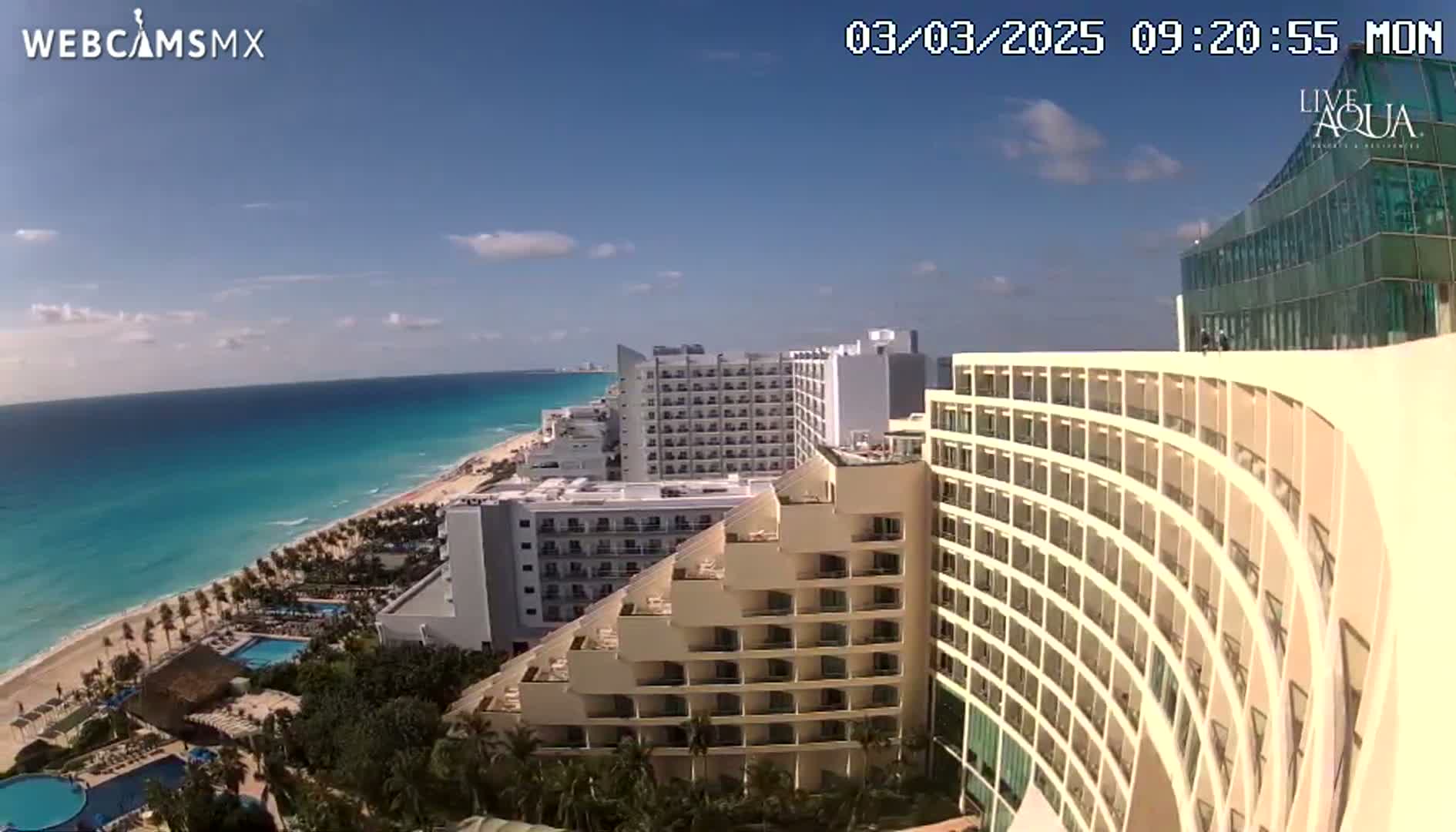 A high-angle view on a sunny day shows a curving hotel overlooking a beach and ocean, with other hotels and resort buildings in the background.