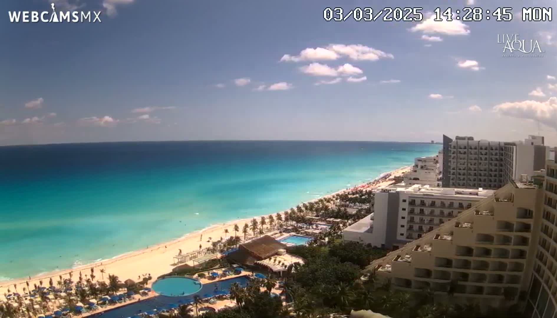 A mostly sunny day shows a long stretch of turquoise ocean lapping a white sand beach lined with hotels and palm trees.