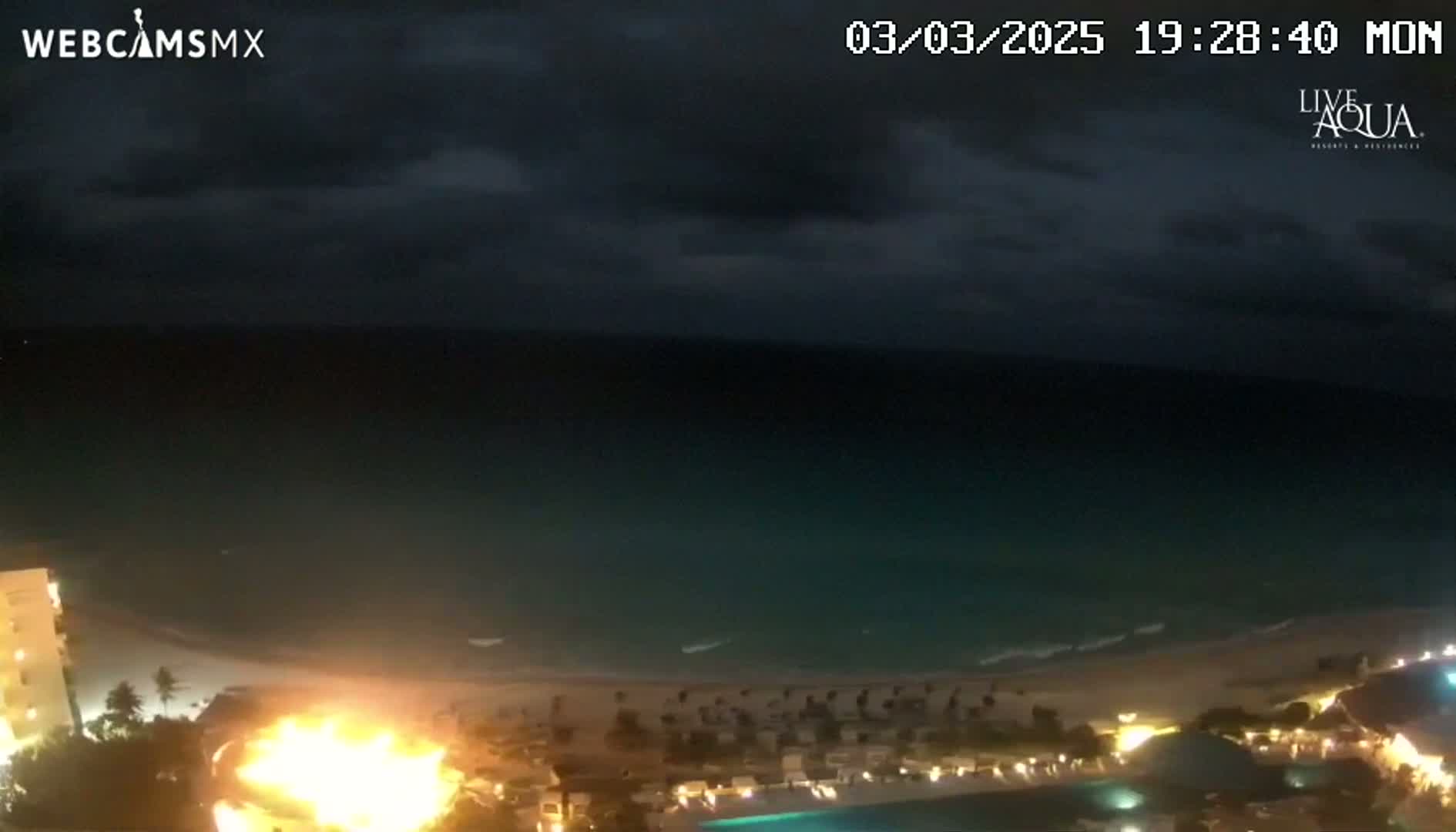 A nighttime view of a beach with a large fire near the shore under a dark, cloudy sky.