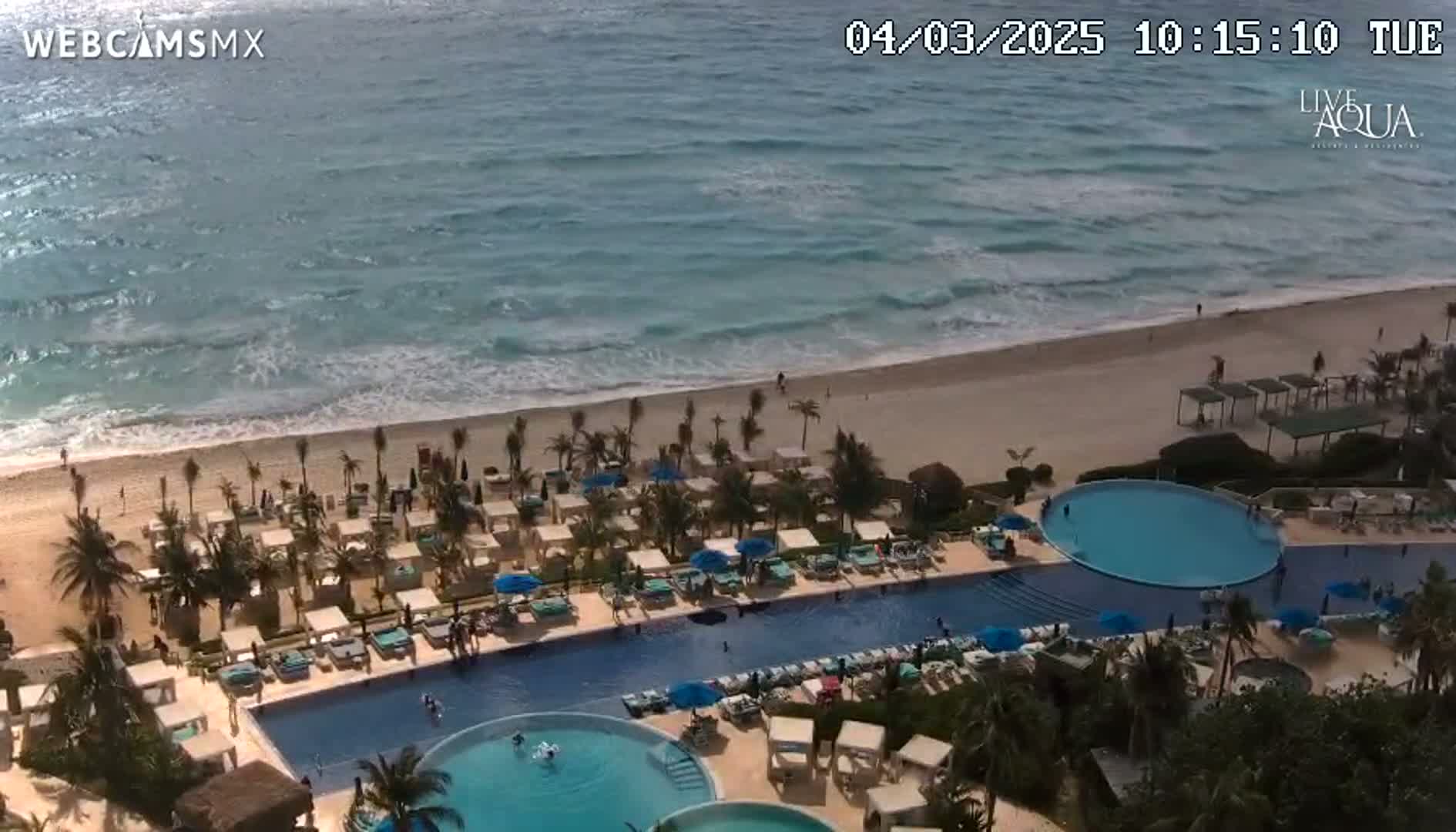 An aerial view shows a resort pool complex adjacent to a beach with moderate waves under sunny skies.