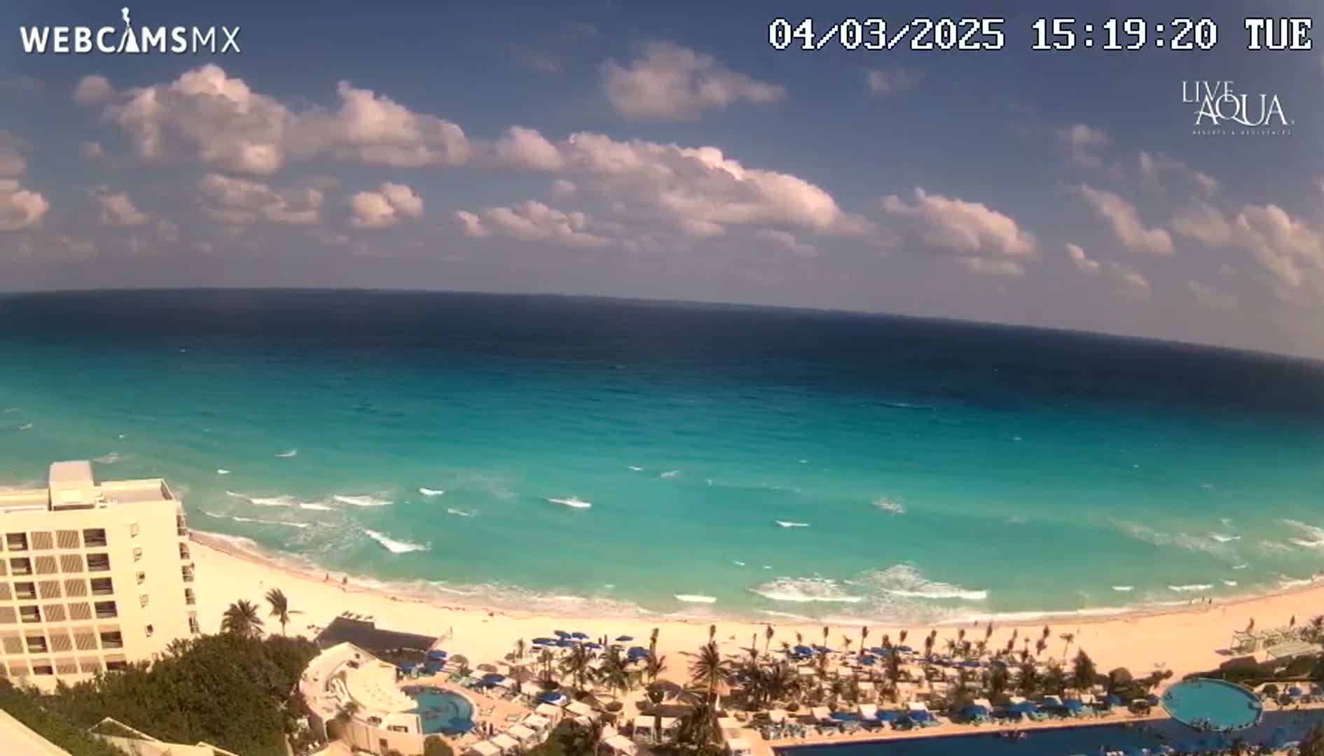 An expansive view of a turquoise ocean, white sandy beach, and resort with palm trees under a partly cloudy sky.