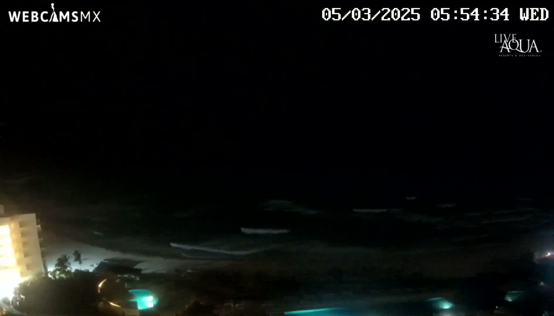 A nighttime view of a dark ocean with small waves lapping a beach, illuminated by artificial lights from buildings and pools nearby.