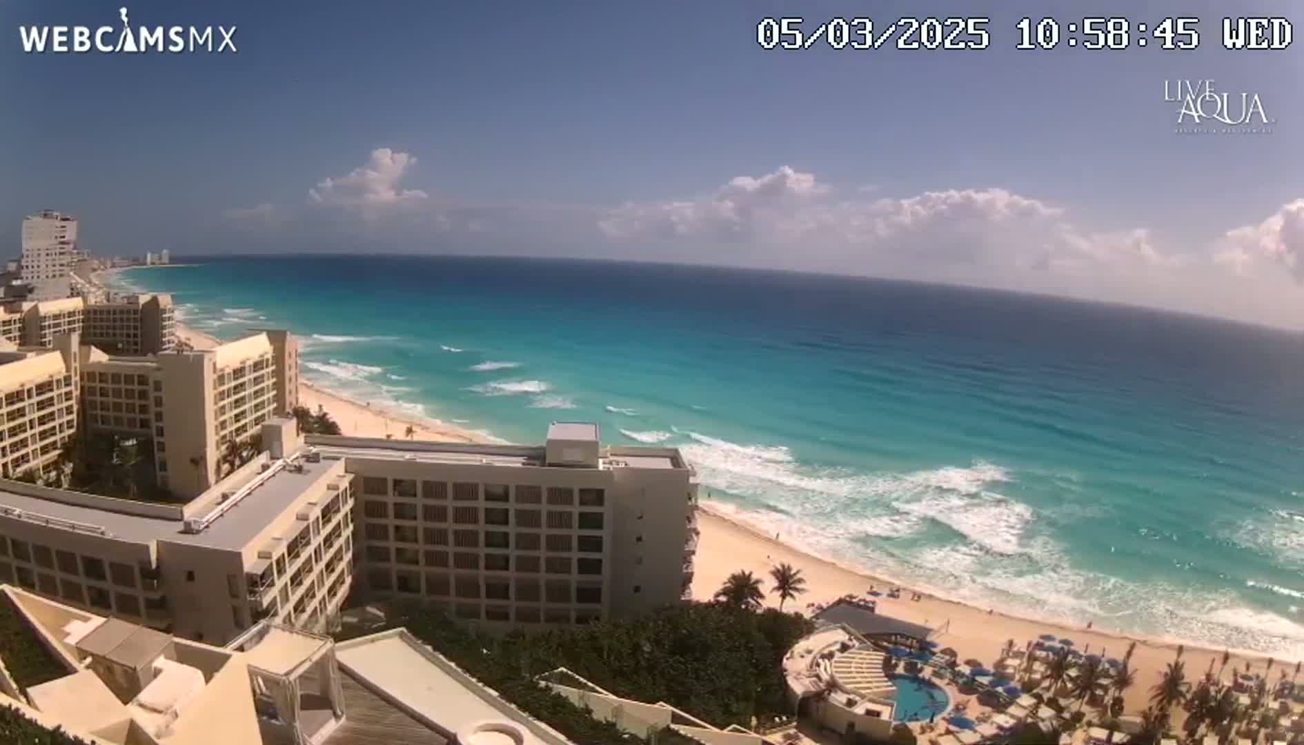 A partly cloudy day reveals a turquoise ocean with waves breaking on a sandy beach lined with hotels and palm trees.