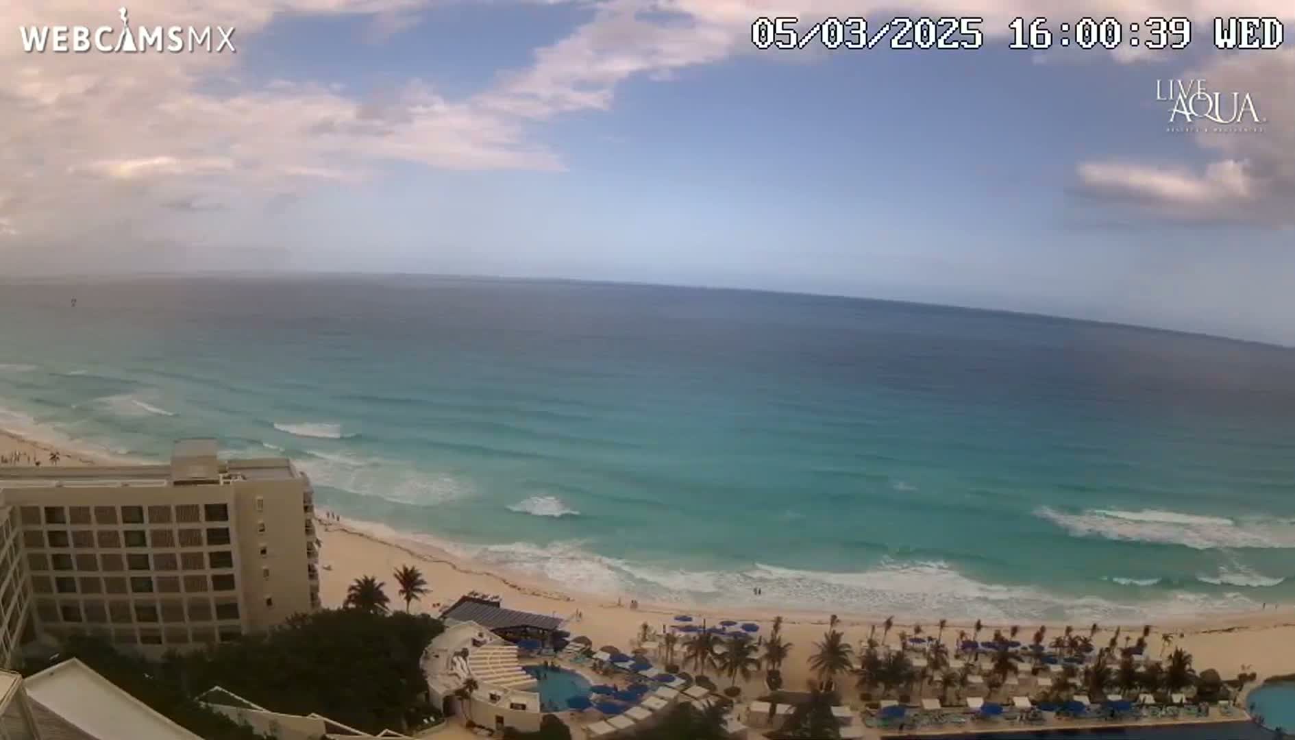 A partly cloudy day shows a beach with turquoise water, gentle waves, and a resort with a pool visible from a high vantage point.