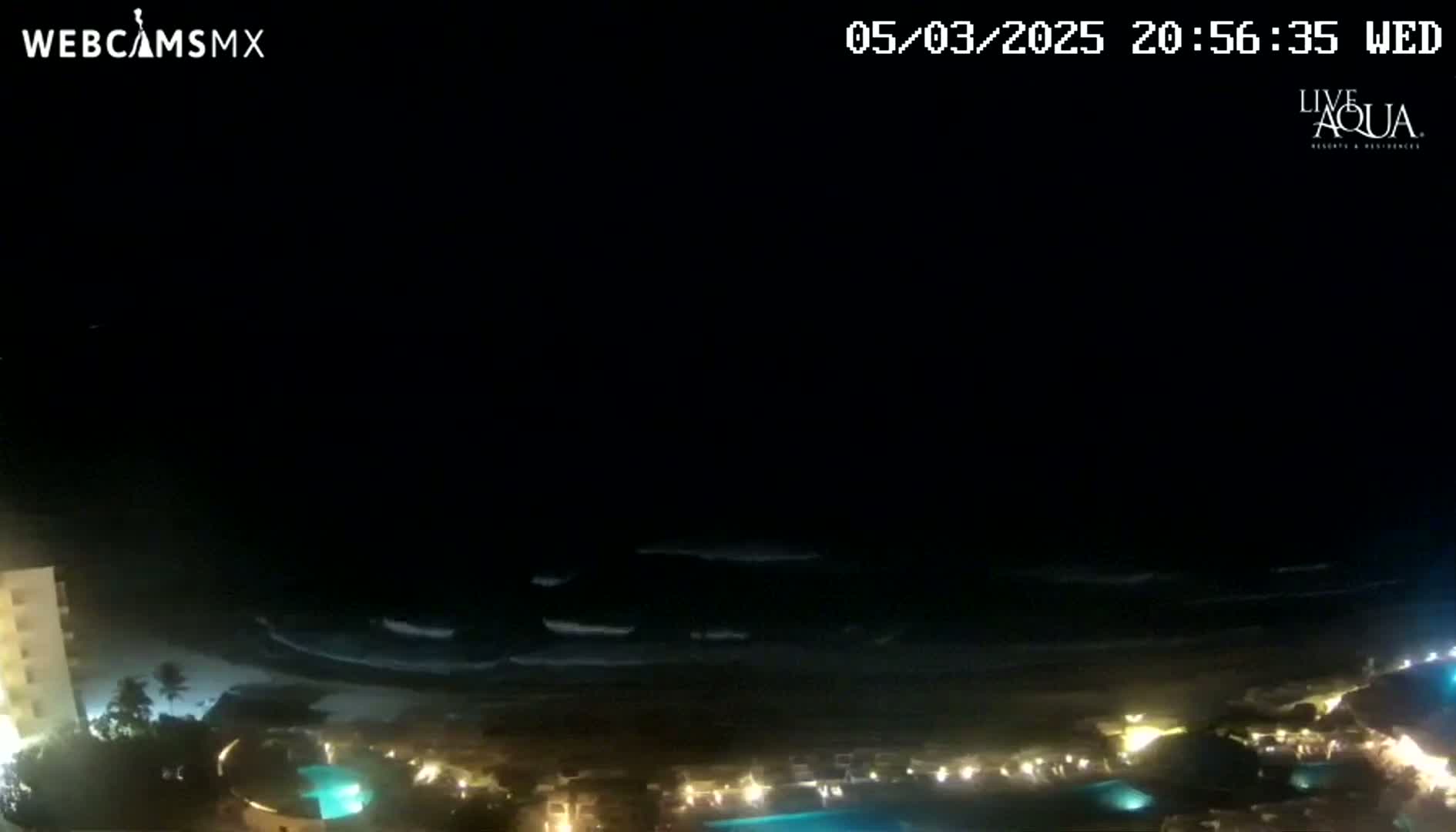 A nighttime view of a coastline shows dark ocean waves lapping at a beach with illuminated buildings and pools along the shore.