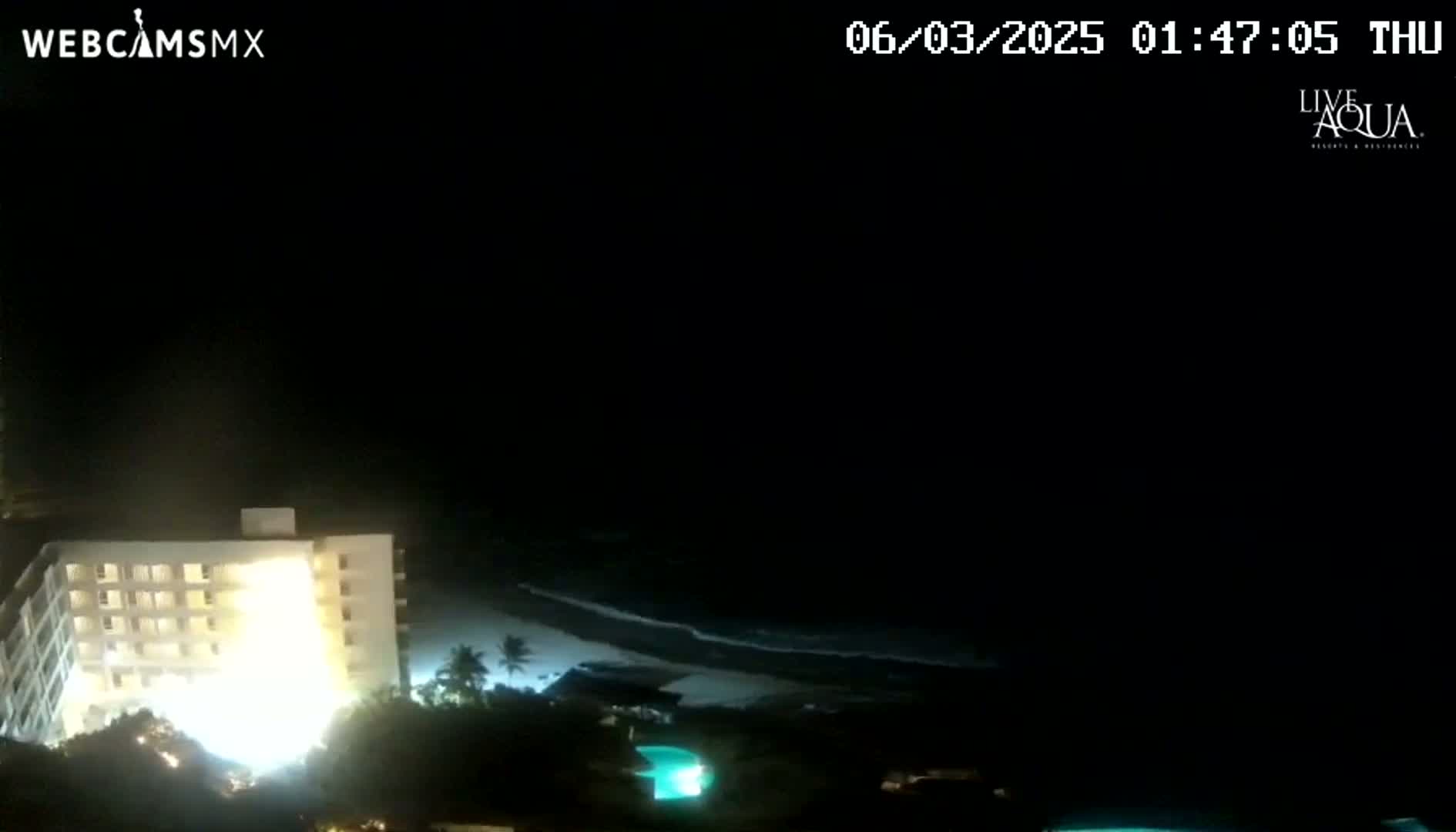 A brightly lit building is seen next to a dark beach with gently lapping waves under a nighttime sky.