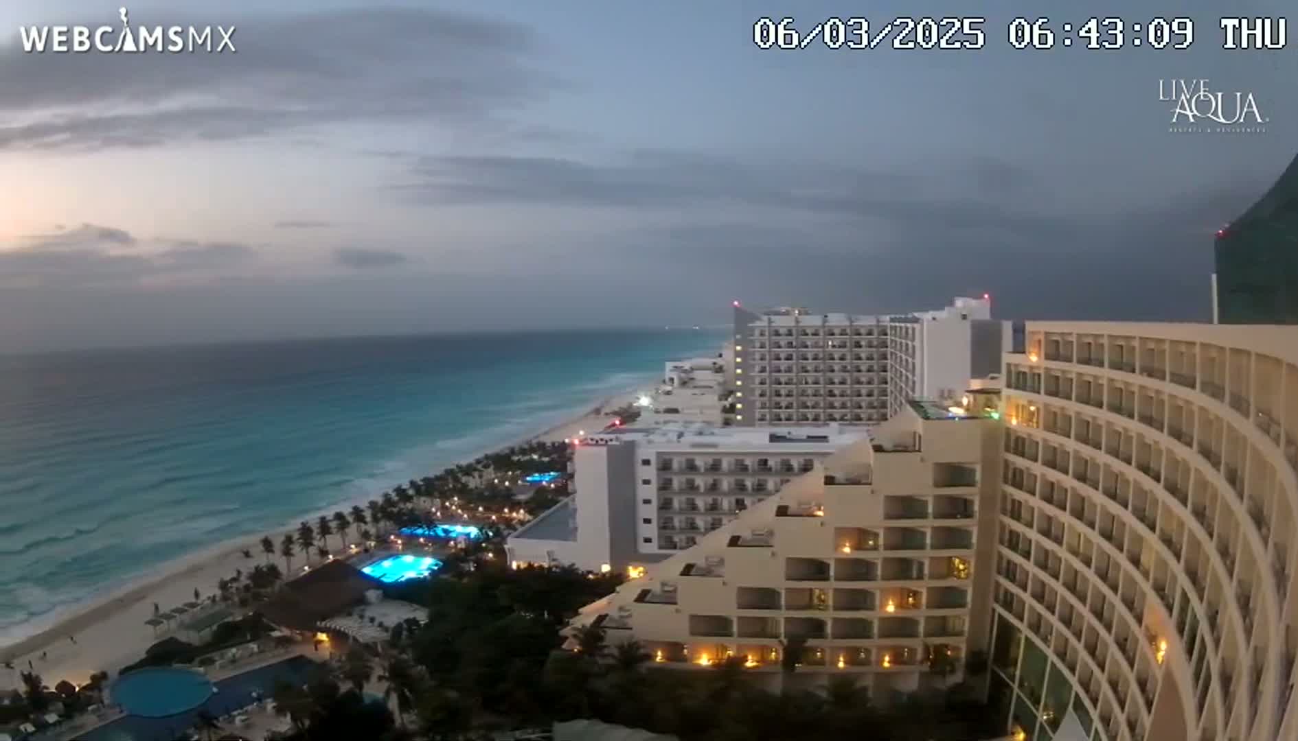 A twilight view of a beachside resort complex with several high-rise hotels, illuminated pools, and a calm ocean under a cloudy sky.
