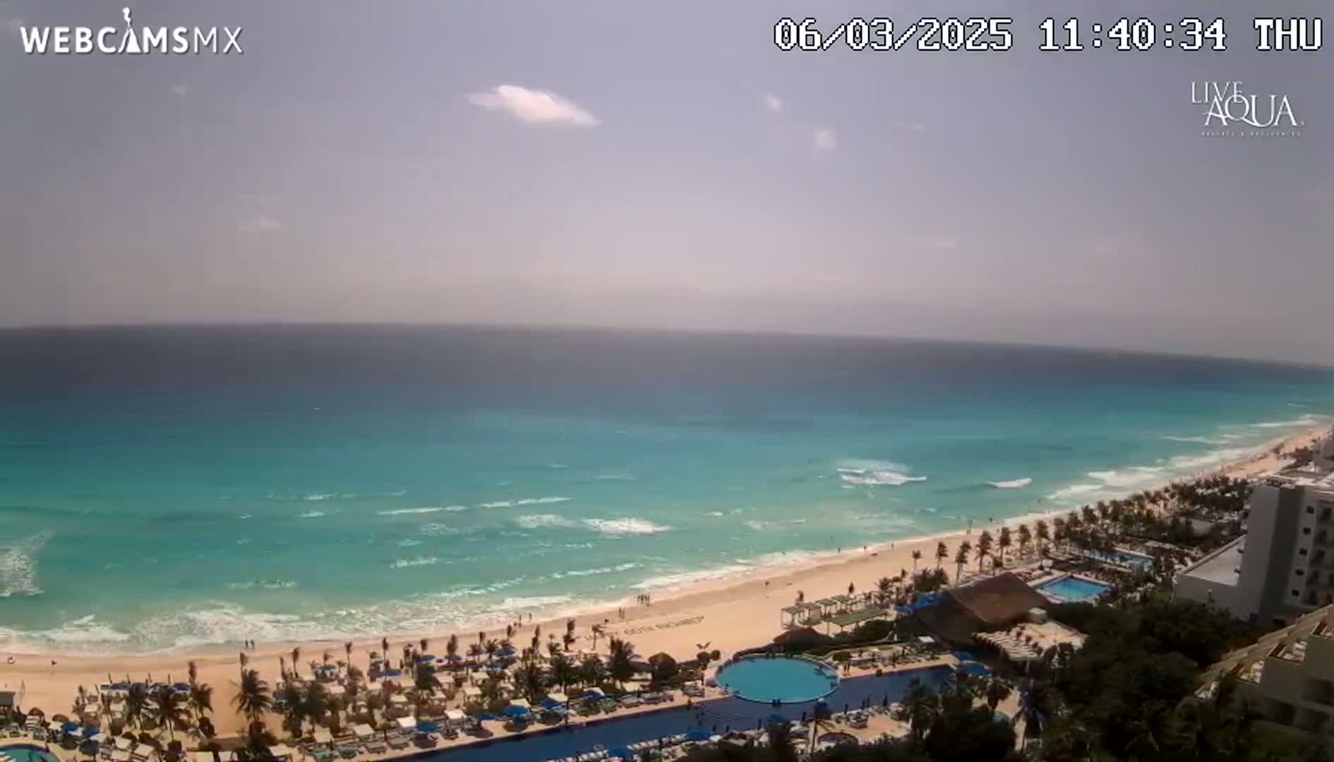 An aerial view shows a mostly sunny beach with turquoise water and gentle waves, overlooking a resort with pools and palm trees.