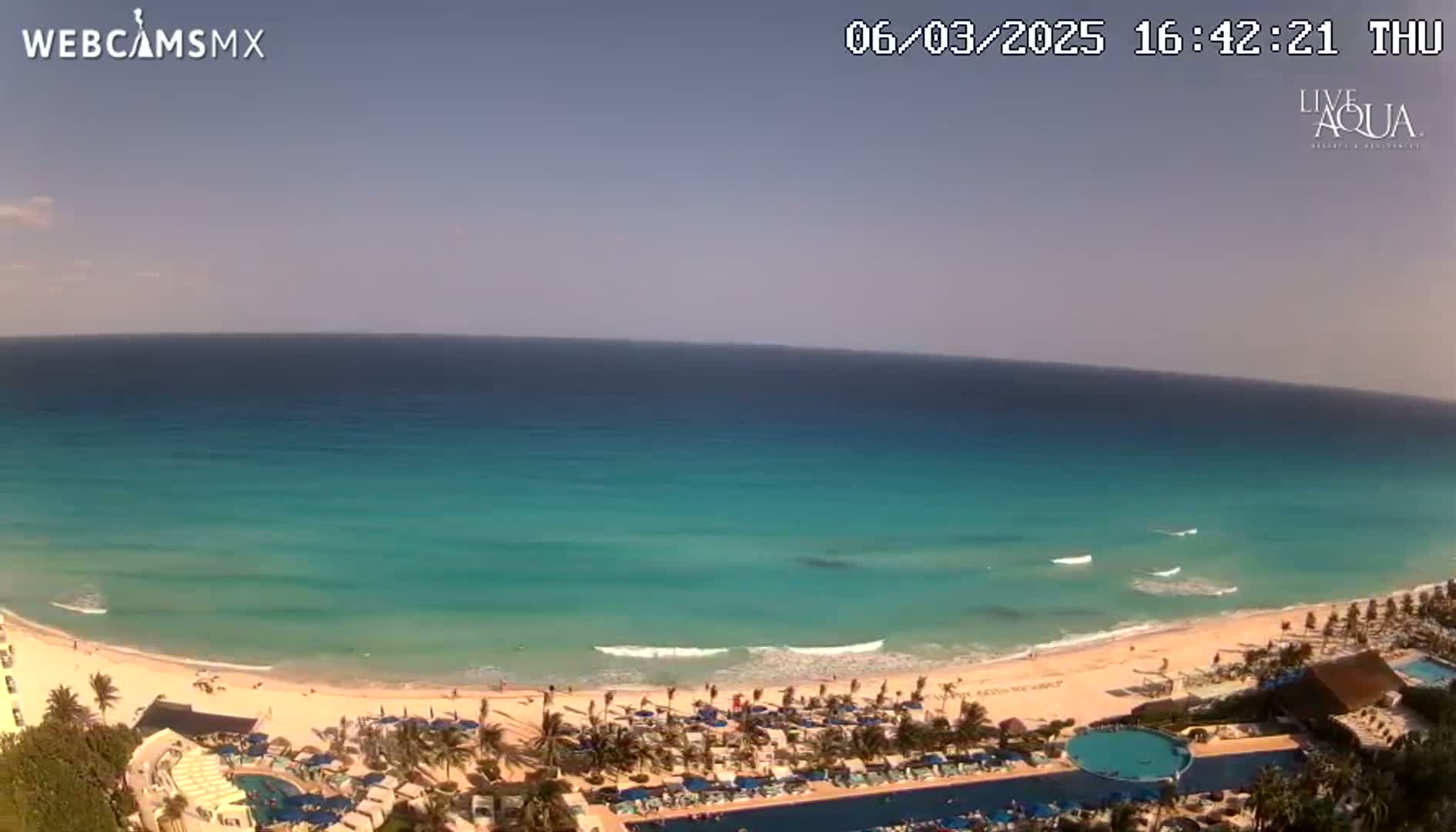 A beach resort with a swimming pool overlooks a calm, turquoise ocean under a clear, sunny sky.