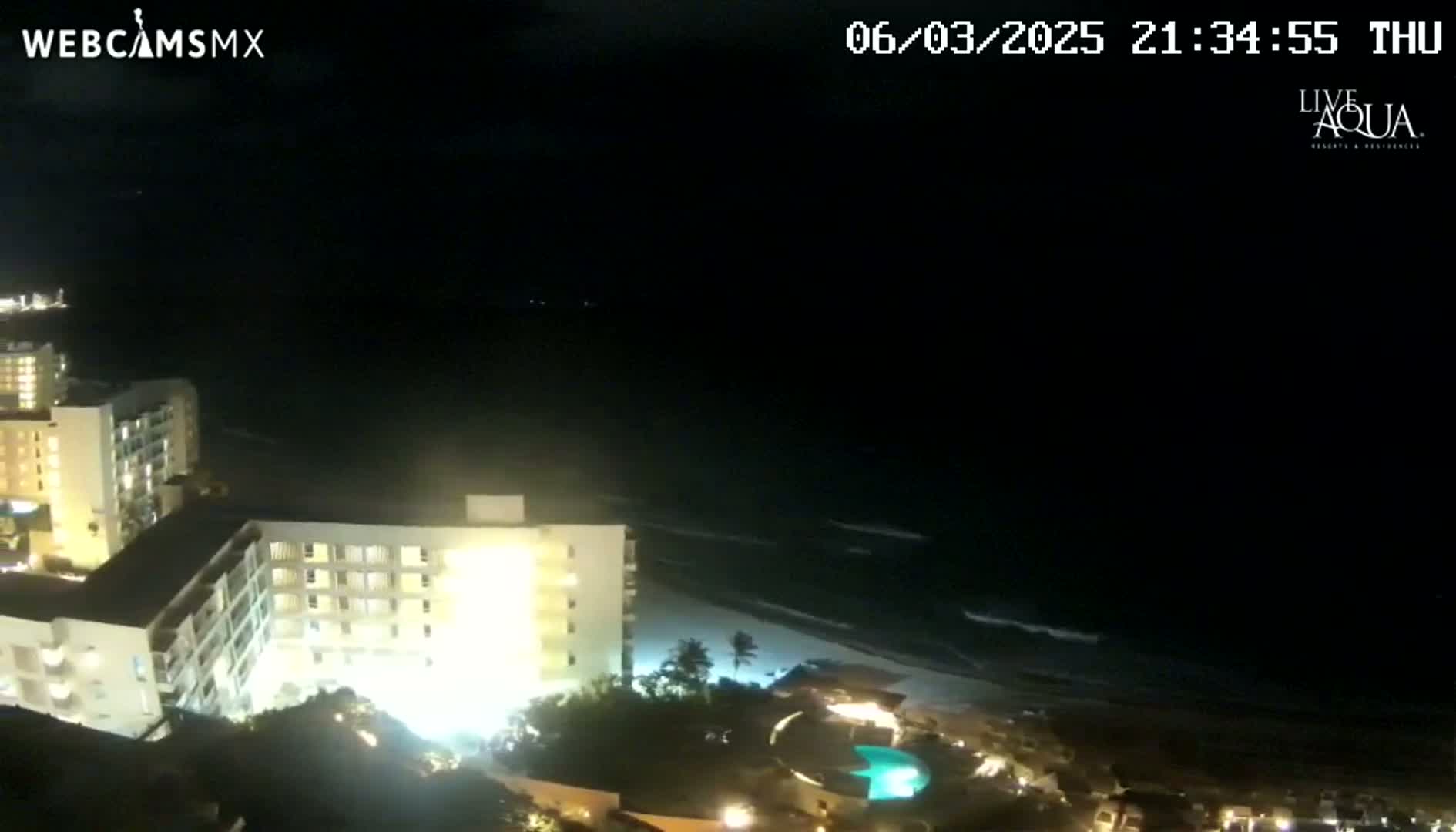 A nighttime view of a beach with gently rolling waves, illuminated by the lights of several brightly lit buildings and a swimming pool.