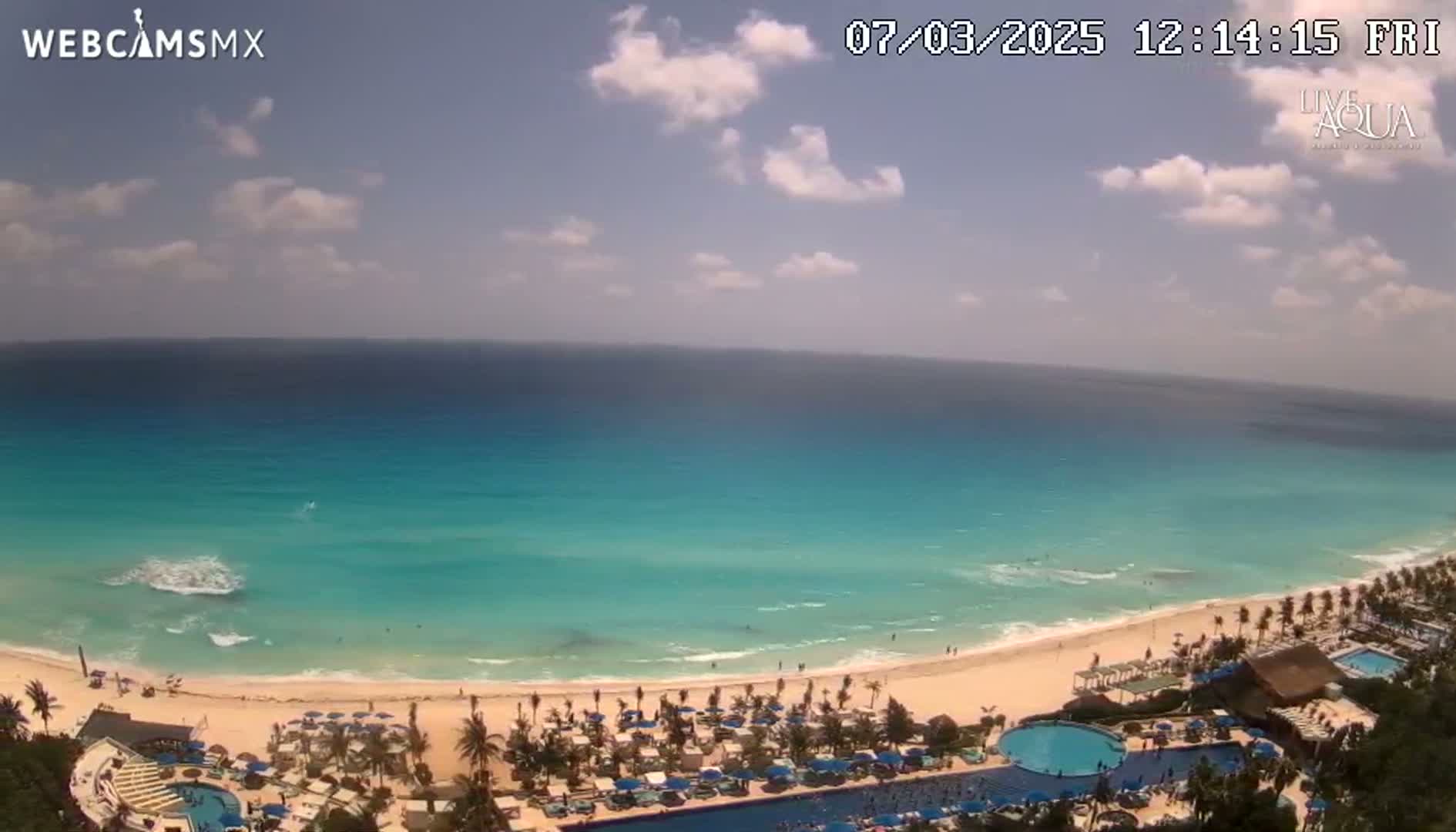 A partly cloudy day shows a beach with turquoise water, a sandy shore lined with palm trees and umbrellas, and a resort with pools.