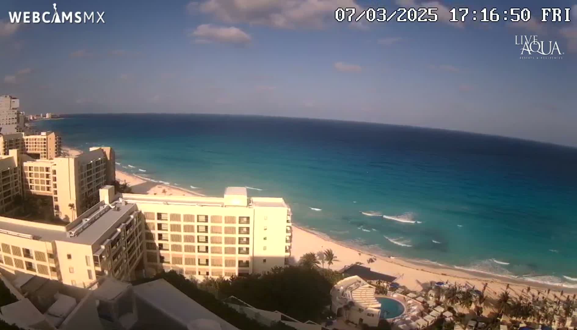 A partly sunny day shows turquoise ocean waves lapping a sandy beach next to several tall beige buildings.