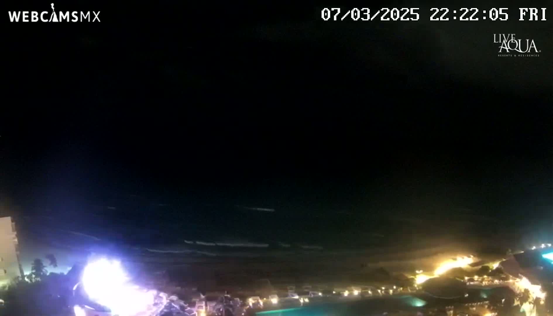 A nighttime view of a beach at a resort, with a bright light possibly from fireworks or lightning near the shore and gentle waves.