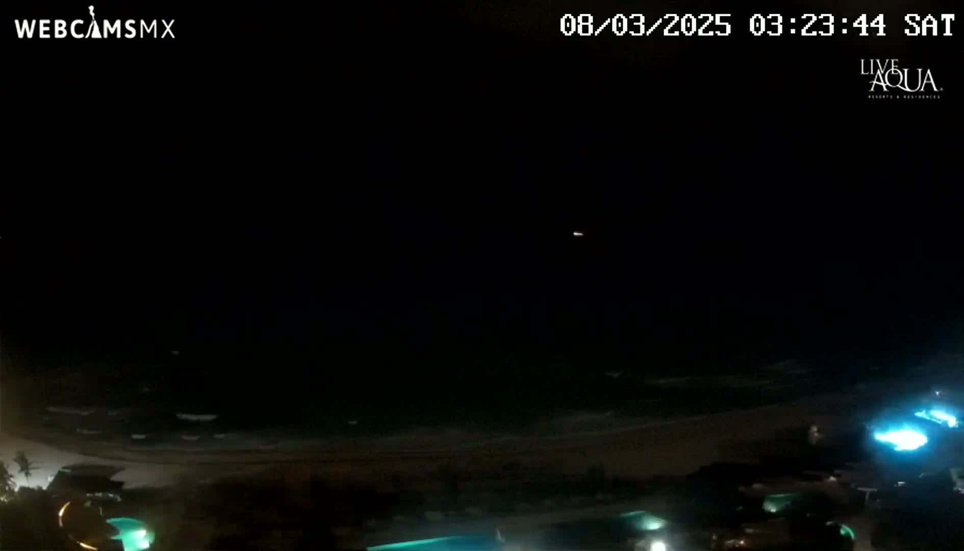 A nighttime view of a dark ocean with gently lapping waves on a beach, featuring illuminated pools and structures in the foreground.