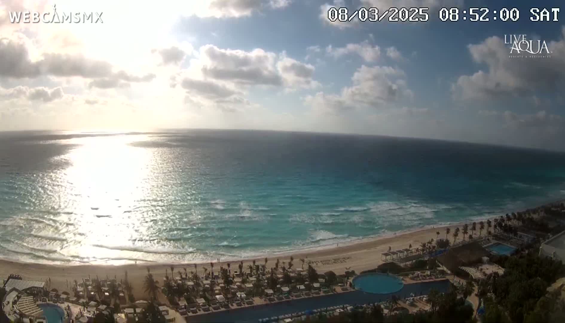 A partly cloudy day reveals a beach resort overlooking a turquoise ocean with gentle waves under bright sunlight.