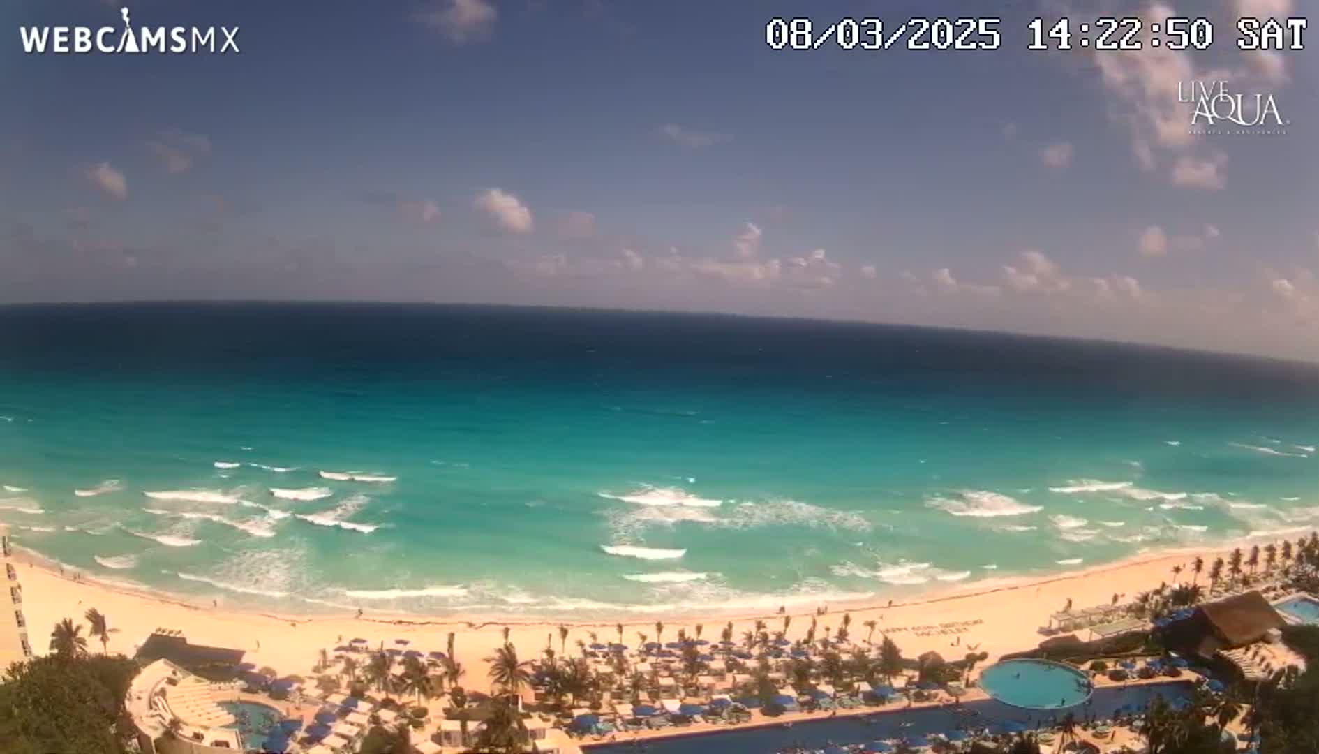 An aerial view shows a beach with turquoise water and small waves under a partly cloudy sky.