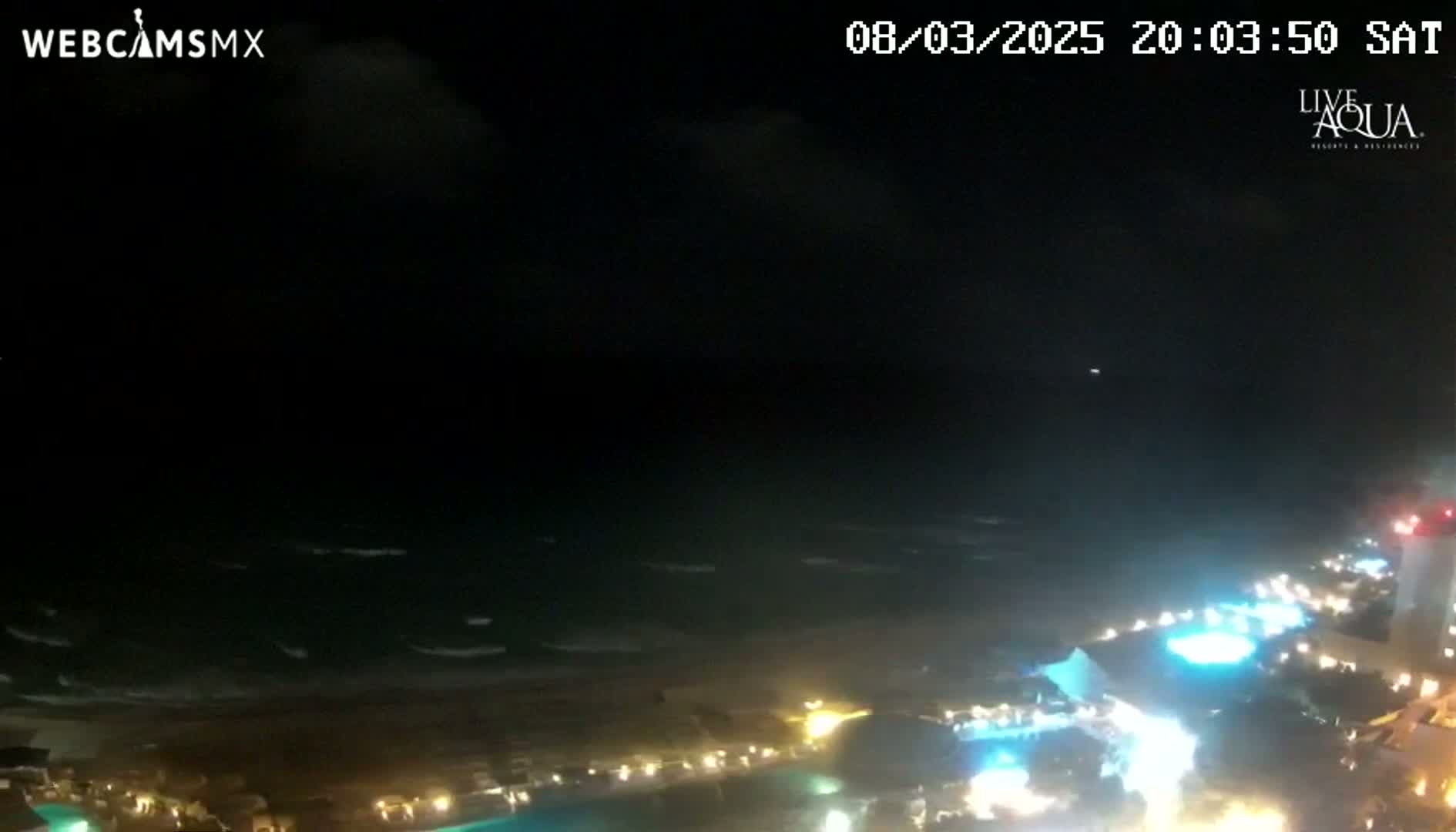 A nighttime coastal scene shows a dark, somewhat cloudy sky over a beach with moderately rough waves, and a shoreline illuminated by lights from buildings and possibly pools.
