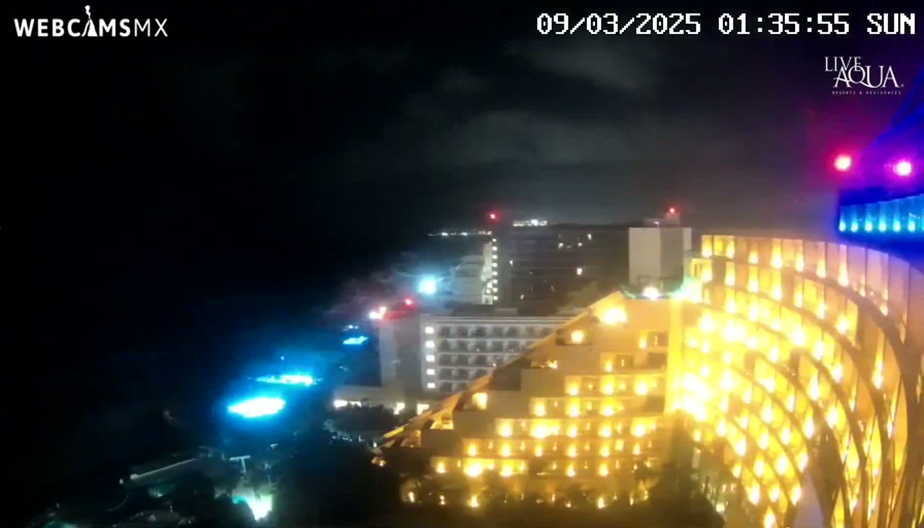 A brightly lit, multi-story hotel is seen at night under a partly cloudy sky, with other buildings and pools visible in the distance.