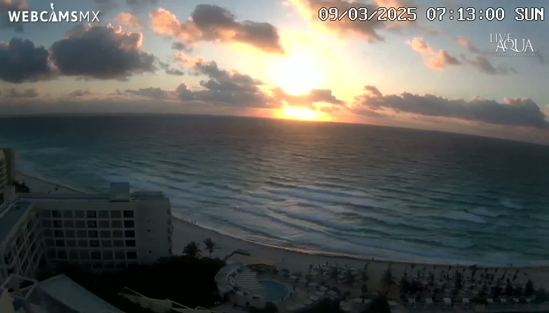 A sunrise over a beach with wavy ocean waters, seen from a high vantage point overlooking a hotel and beach umbrellas.