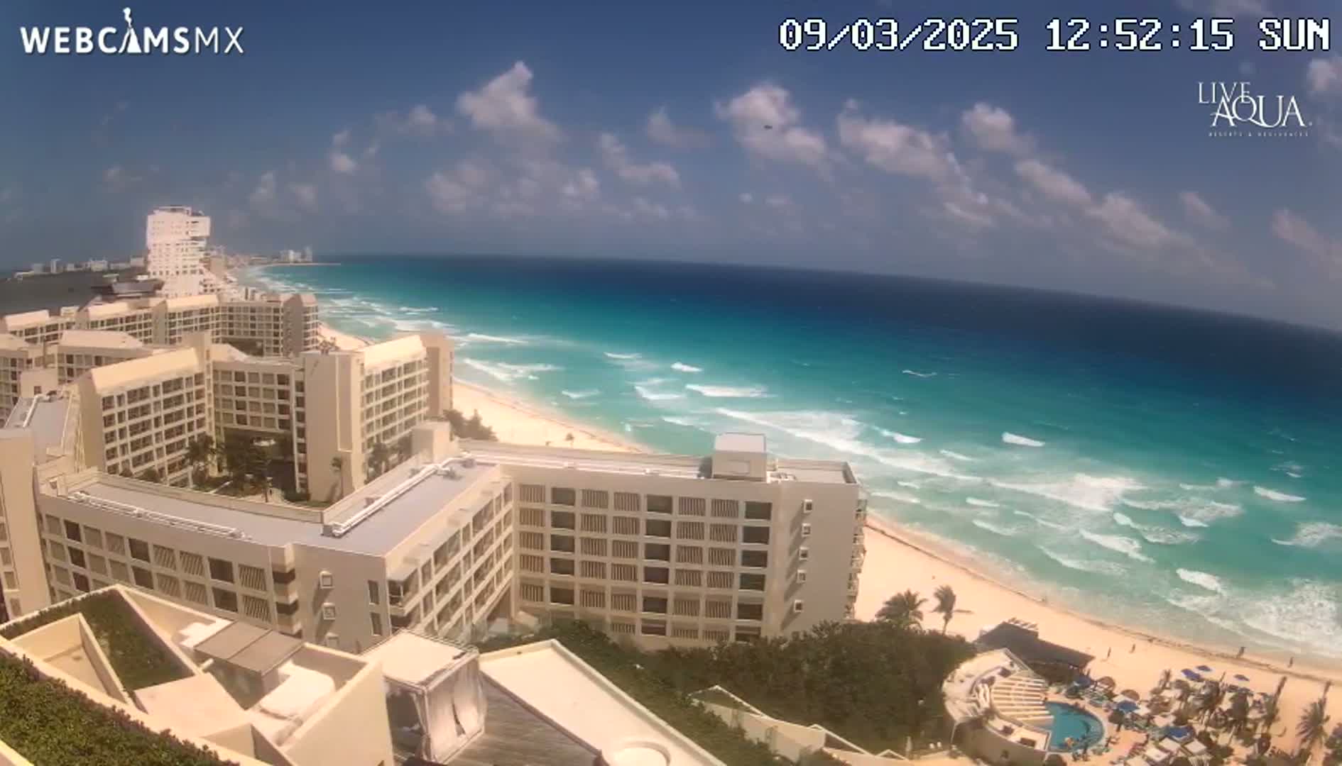 A sunny day shows a beach with turquoise water and waves lapping a sandy shore, lined with multiple large hotels and resort buildings.