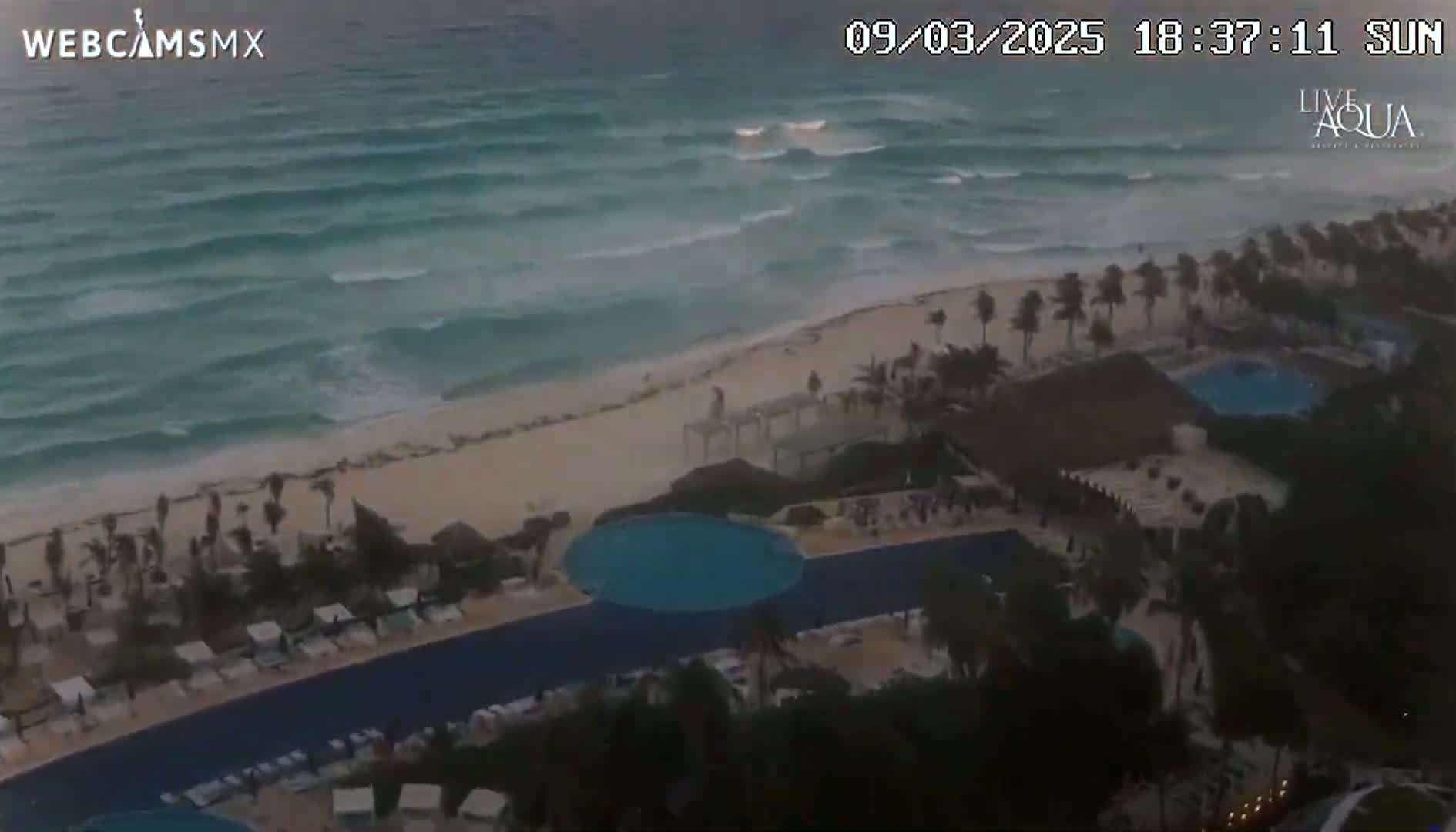 An aerial view shows a beach with moderate waves at sunset, adjacent to a resort with multiple pools and palm trees.