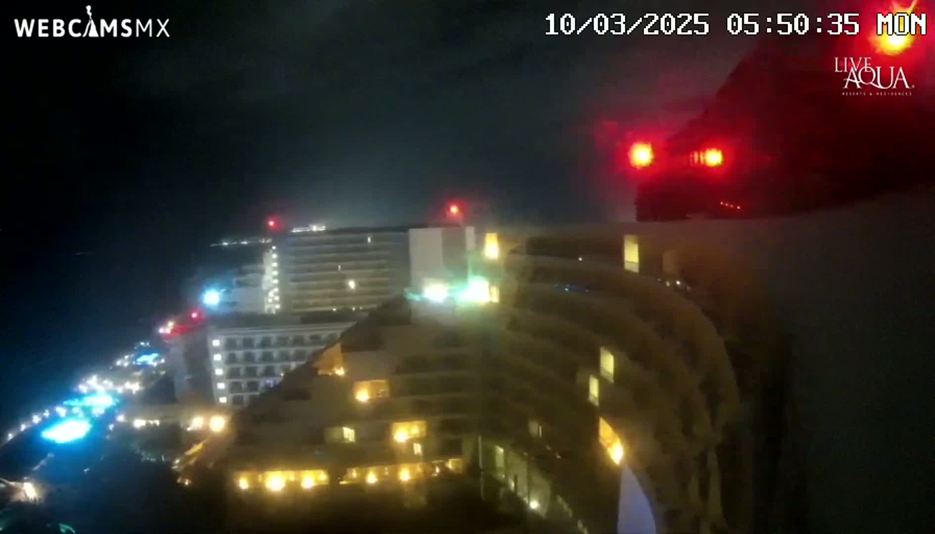 A nighttime, high-angle view shows brightly lit hotel buildings along a coastline, with a vehicle's red taillights visible in the upper right corner.