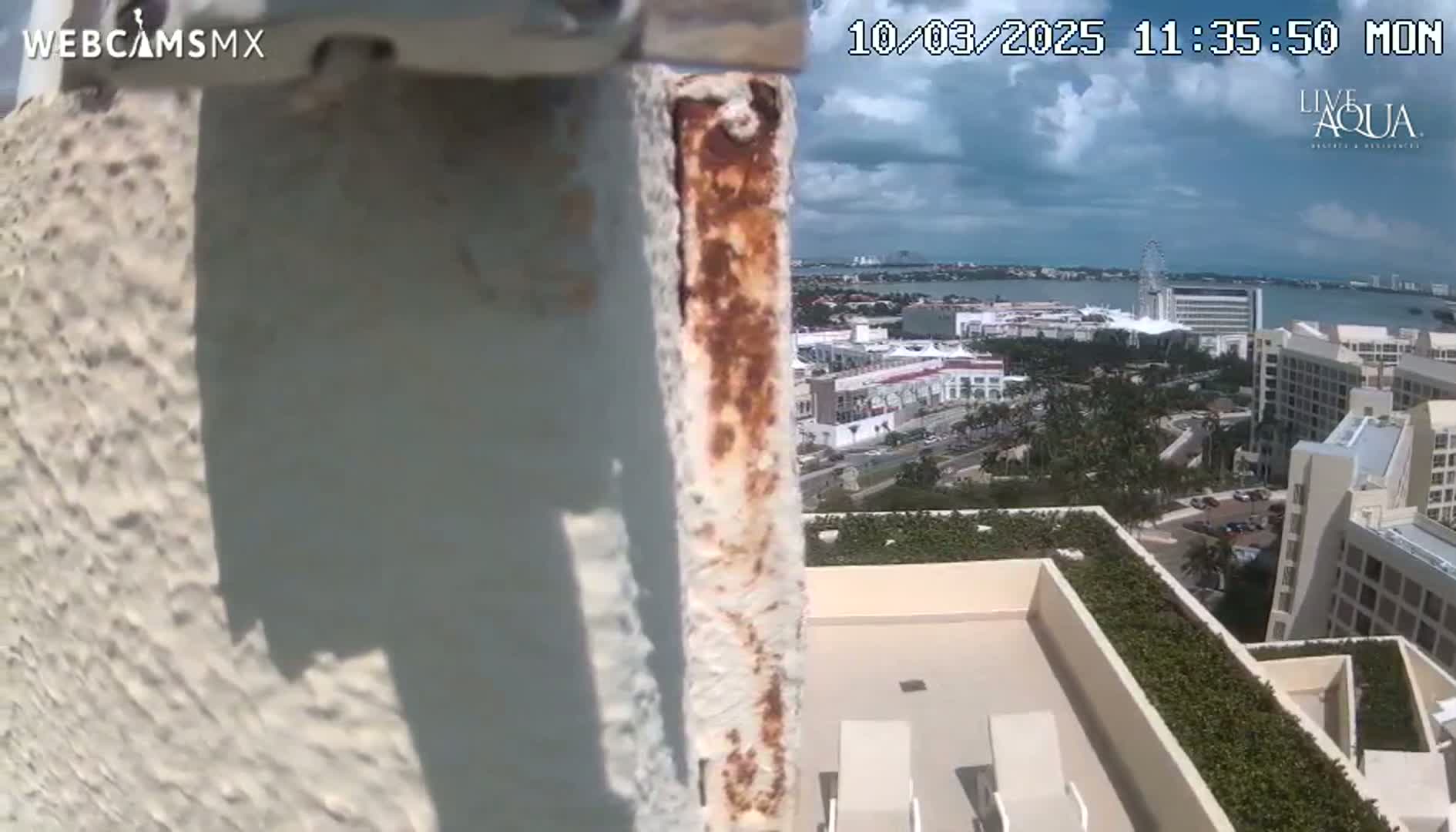 A rusty metal pole is in the foreground, with a partly cloudy sky and a view of a resort town and body of water in the background.