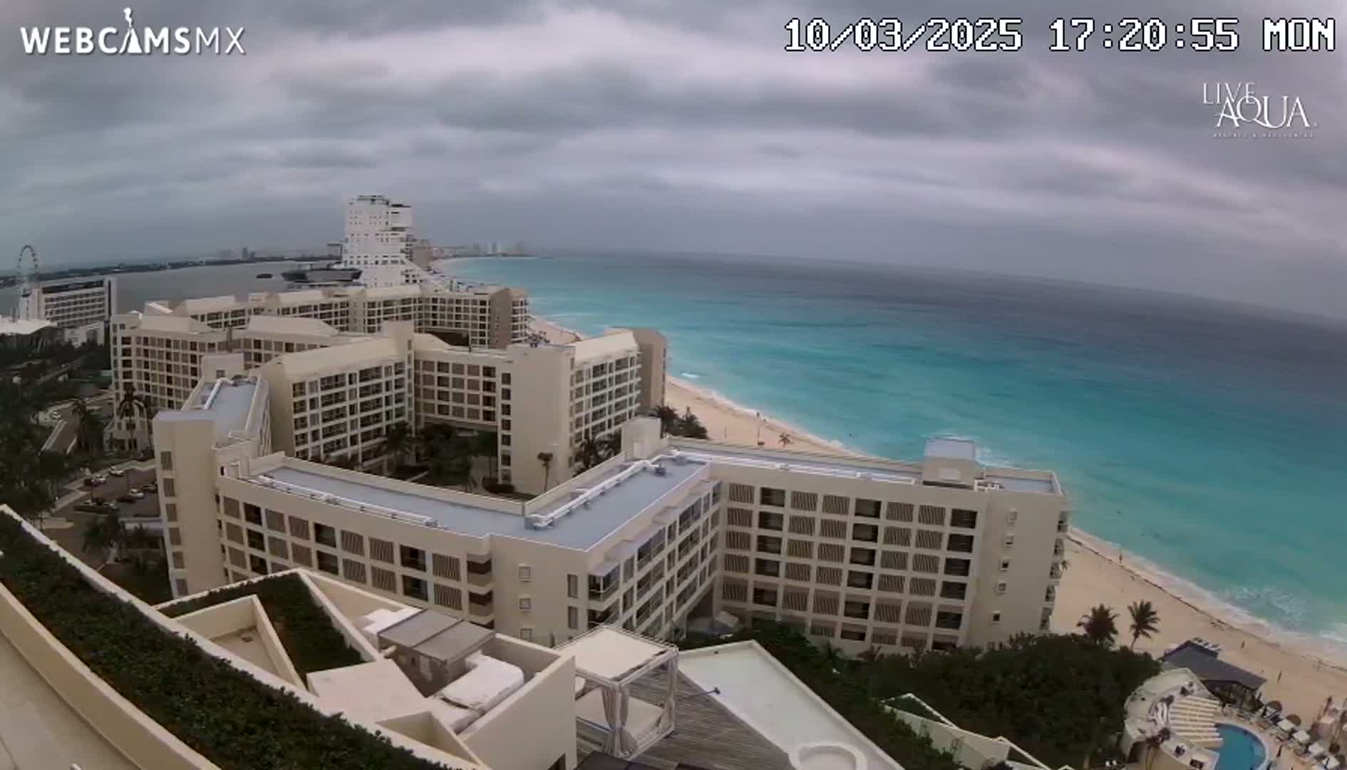 An aerial view shows a beachside resort complex under an overcast sky, with turquoise water visible in the background.