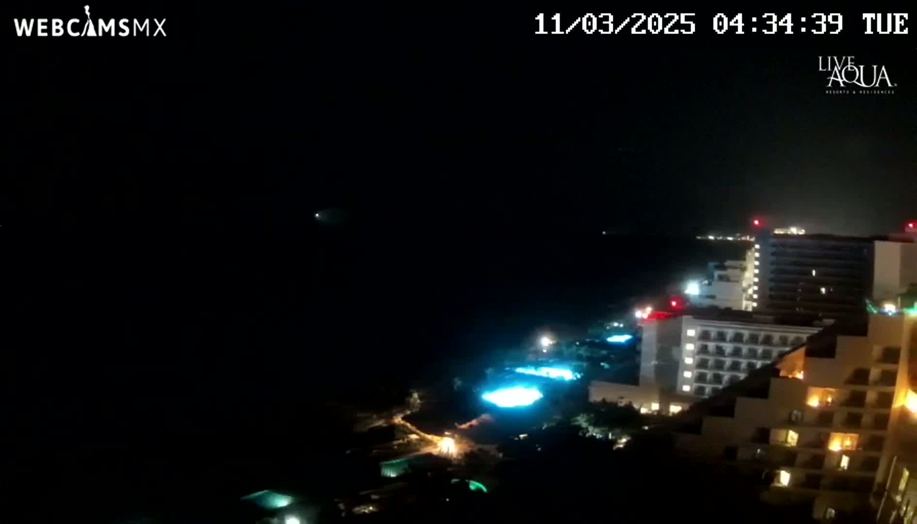 A nighttime coastal view shows illuminated hotels and pools along a dark ocean under a clear, dark sky.