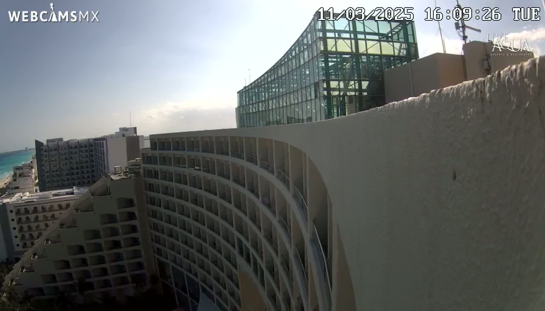 A curved, multi-story hotel overlooks a beach and ocean under a mostly sunny sky.
