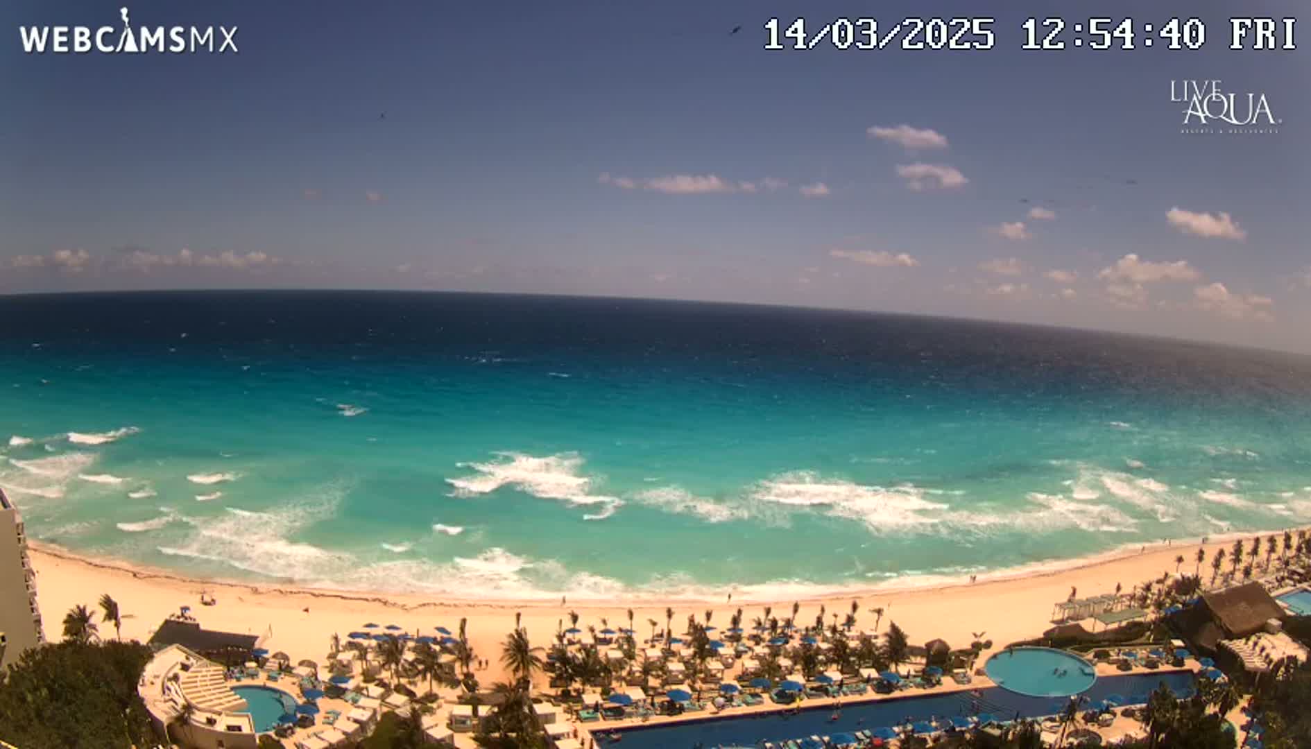 An expansive view of a beach resort with a turquoise ocean under partly sunny skies and moderate waves.