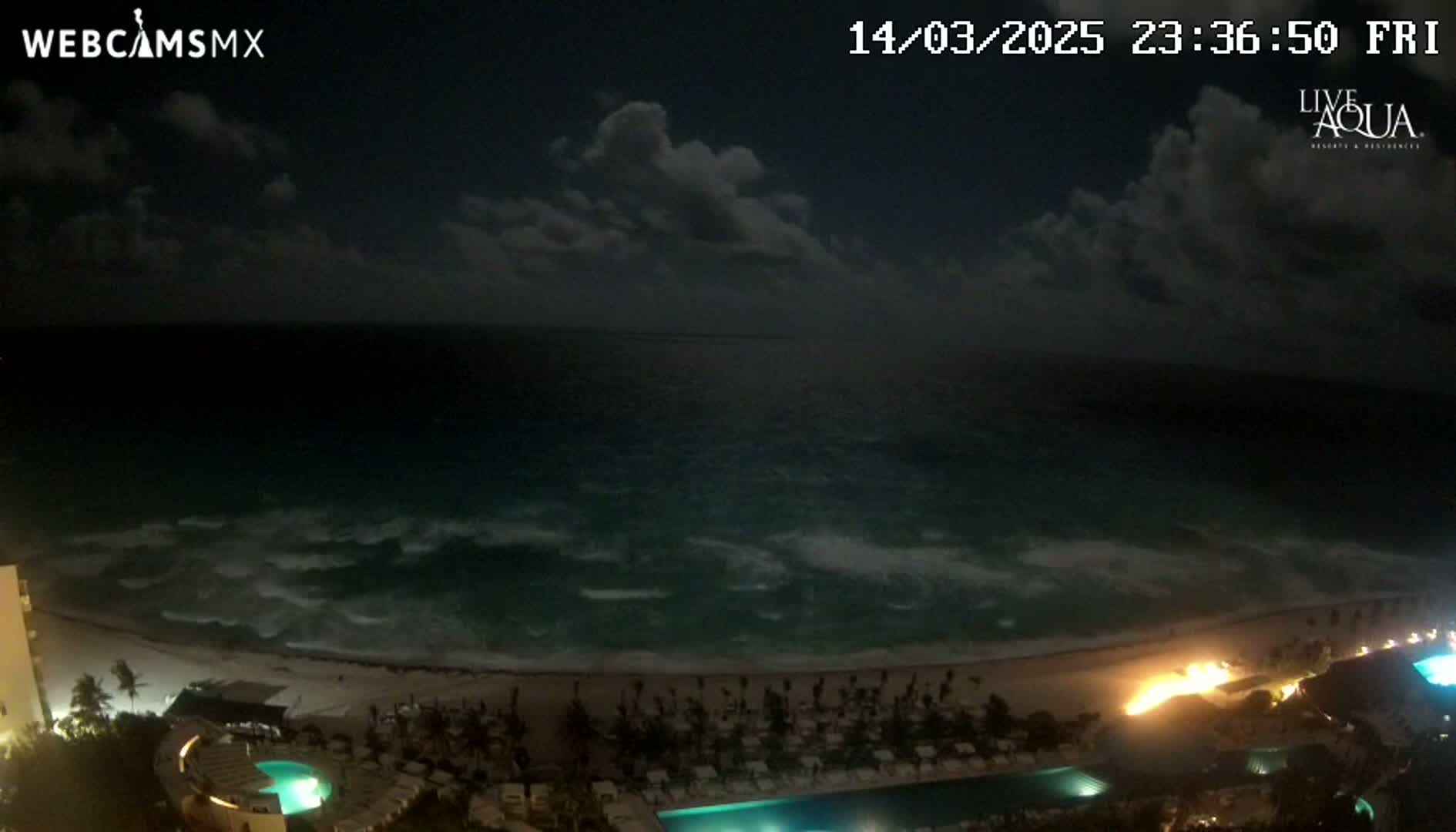 A nighttime view shows a beach with moderately wavy ocean under a partly cloudy sky, featuring a resort with pools and lights along the shore.