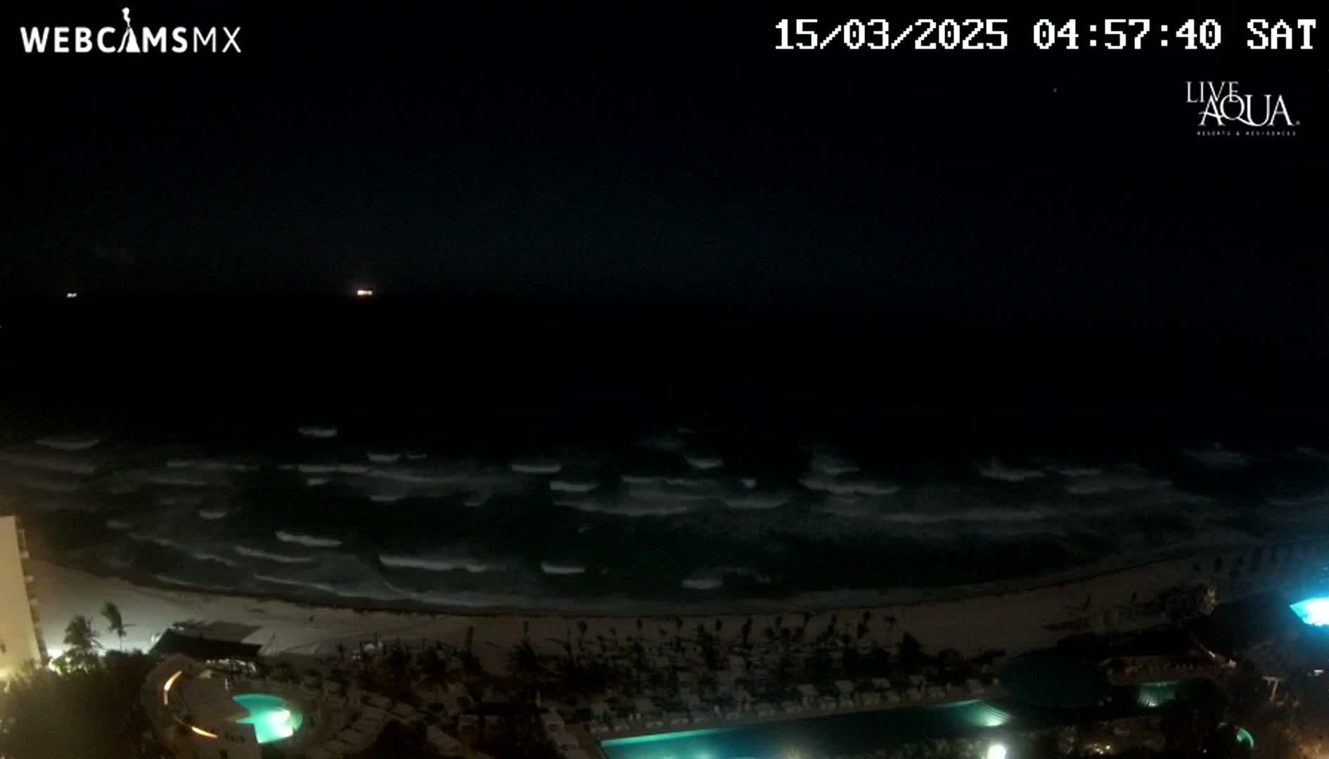 A nighttime view of a beach with moderate waves, showing a resort with pools and surrounding vegetation under a dark sky.