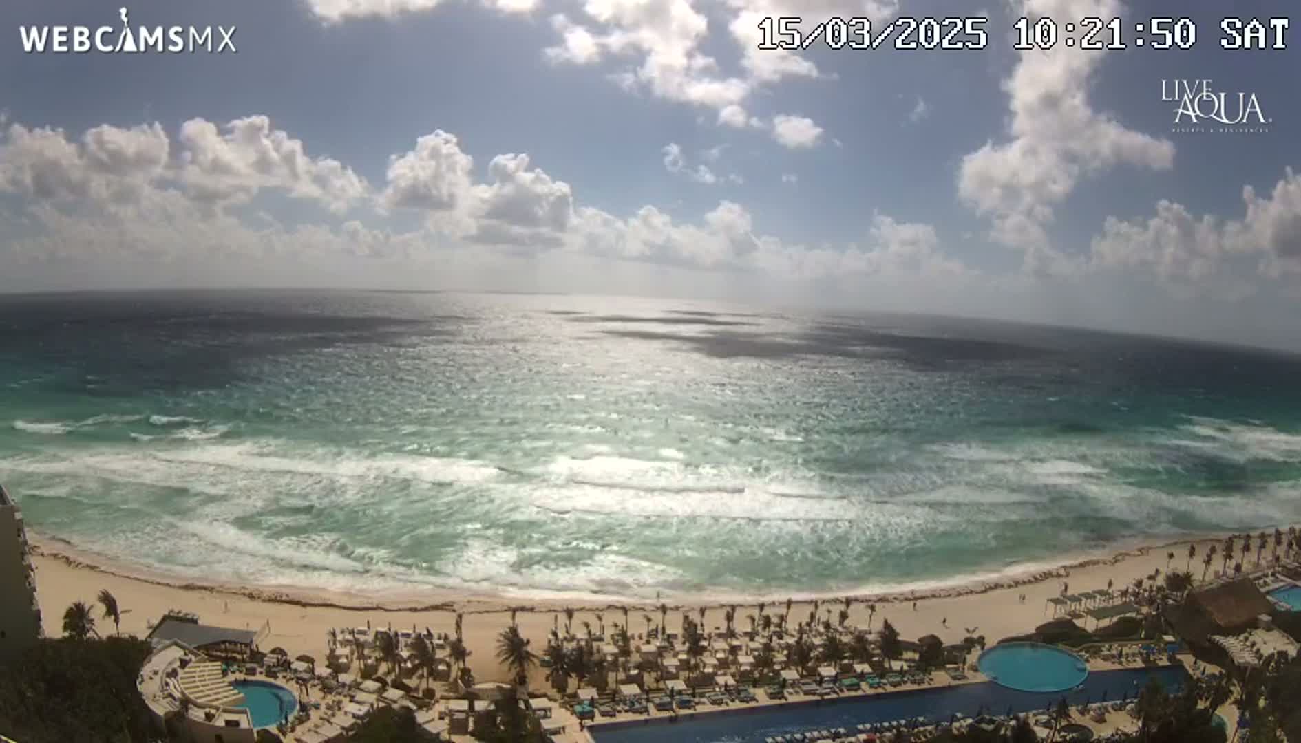 A partly cloudy day shows a beach resort overlooking a turquoise ocean with moderate waves.