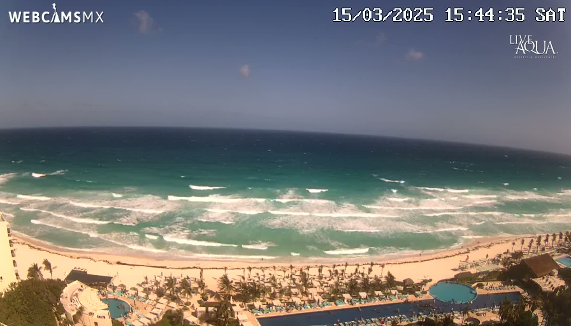 An aerial view shows a beach with turquoise water and moderate waves under a clear blue sky.