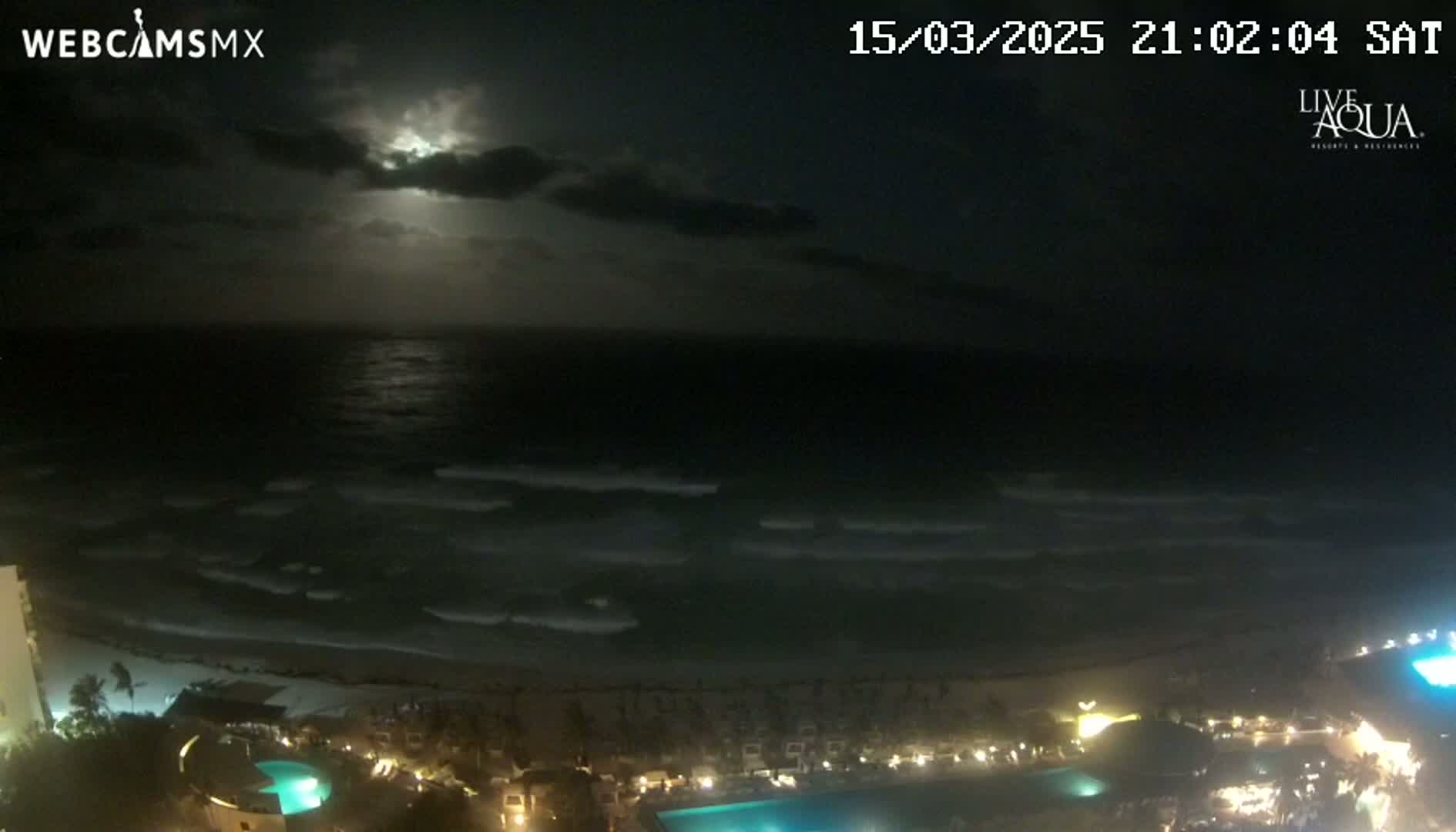 A nighttime view of a moonlit ocean with gentle waves lapping a beach, overlooking a resort with illuminated pools and buildings under a partly cloudy sky.