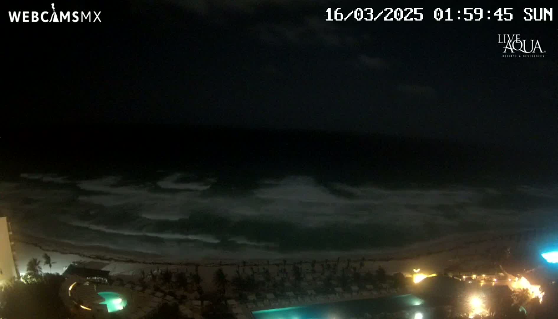 A nighttime view of a beach with waves crashing on the shore, illuminated by the lights of a nearby resort.