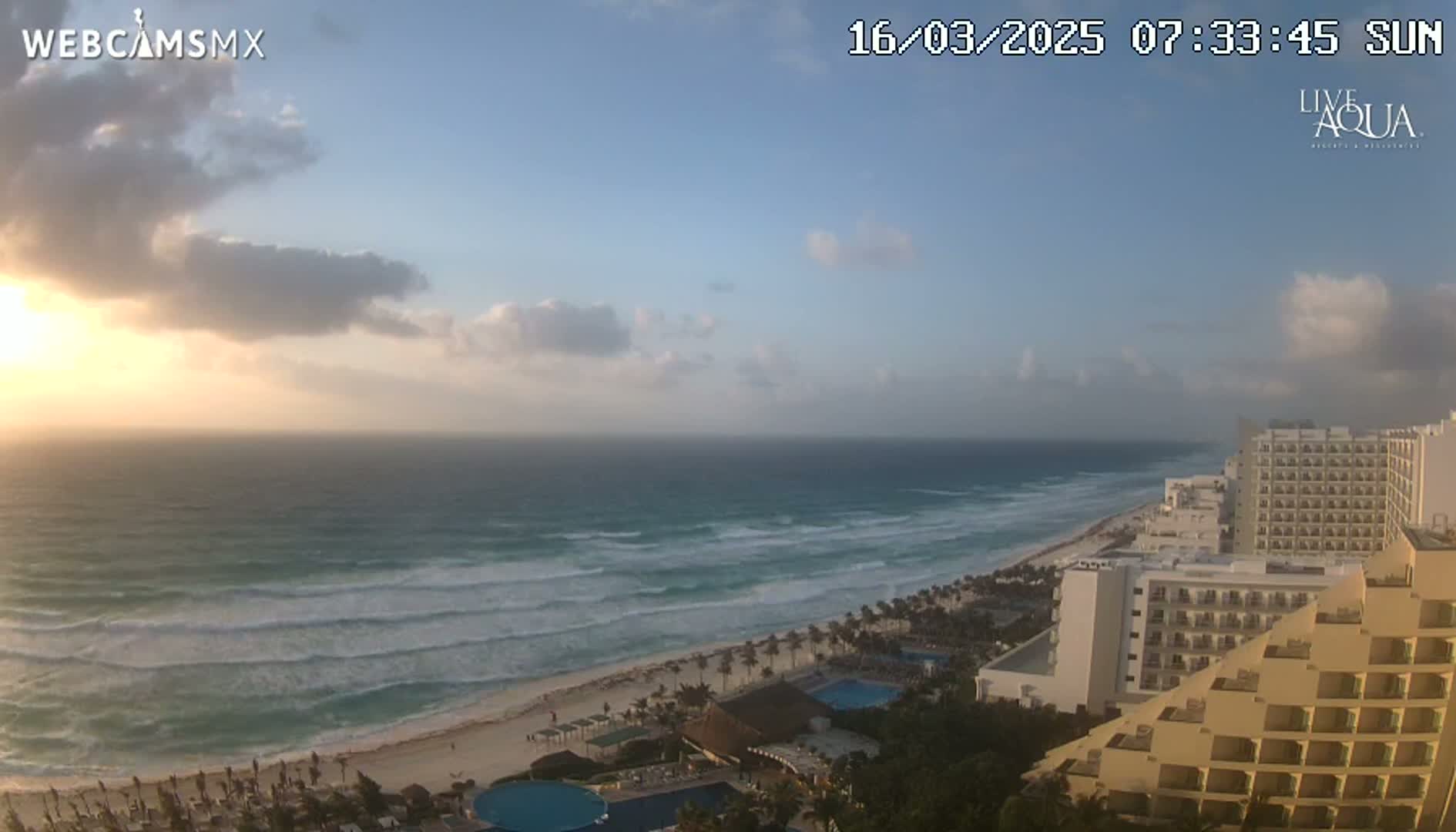 A partly cloudy sunrise illuminates a beach with moderate waves and several high-rise hotels.