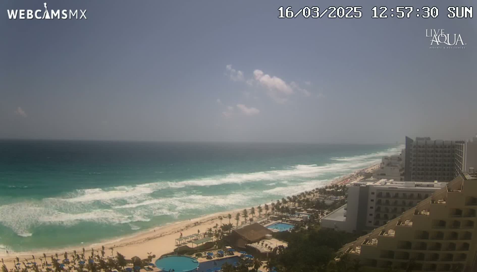 A mostly sunny day shows a beach with turquoise water, waves breaking on the shore, and hotels along the coast.