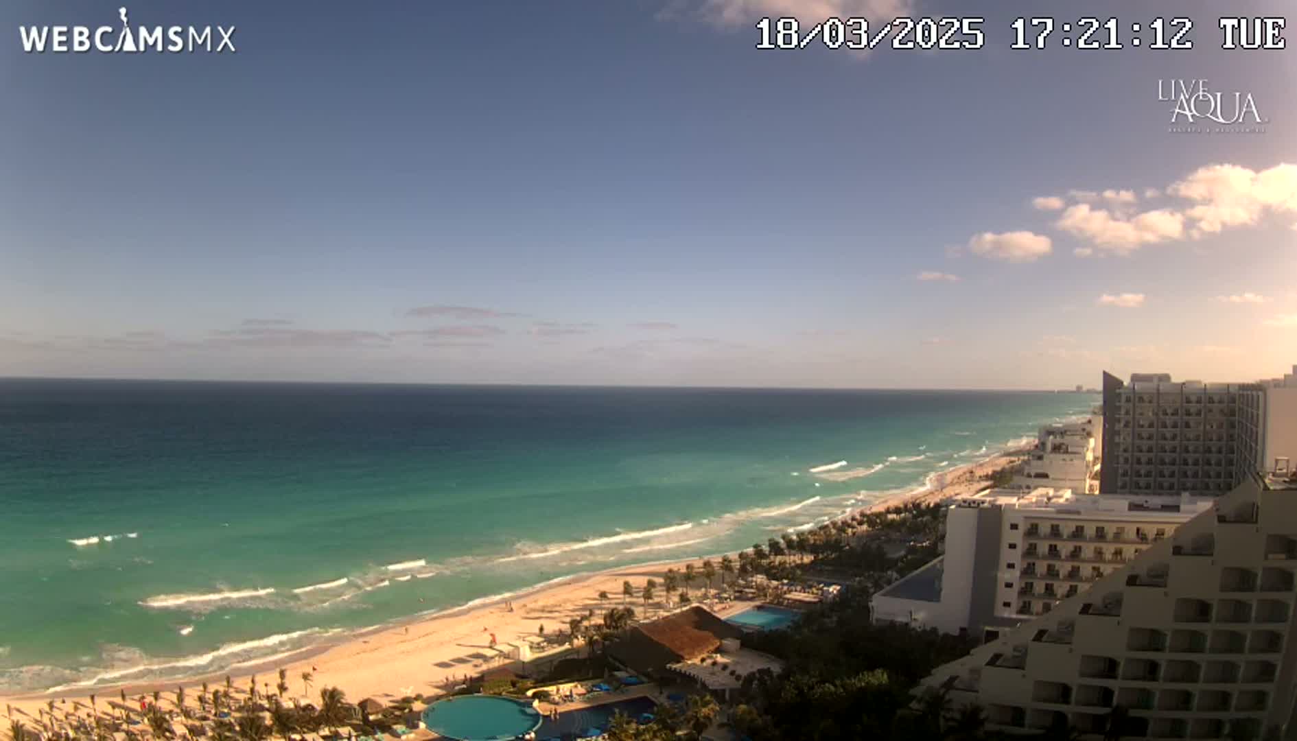 A mostly sunny day reveals a long stretch of sandy beach and turquoise ocean with gentle waves, overlooked by several high-rise hotels.