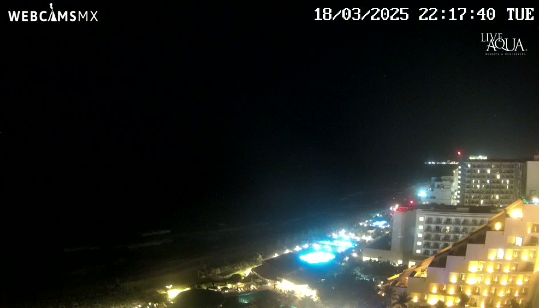 A nighttime, high-angle view shows a resort town's brightly lit buildings along a dark ocean shoreline under a clear, starlit sky.