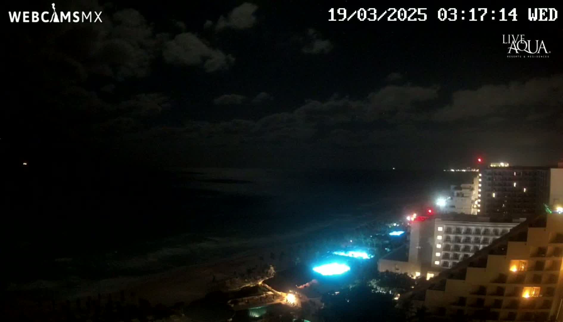 A nighttime view of a coastline shows a dark ocean under a partly cloudy sky, with illuminated hotels and pools along the shore.