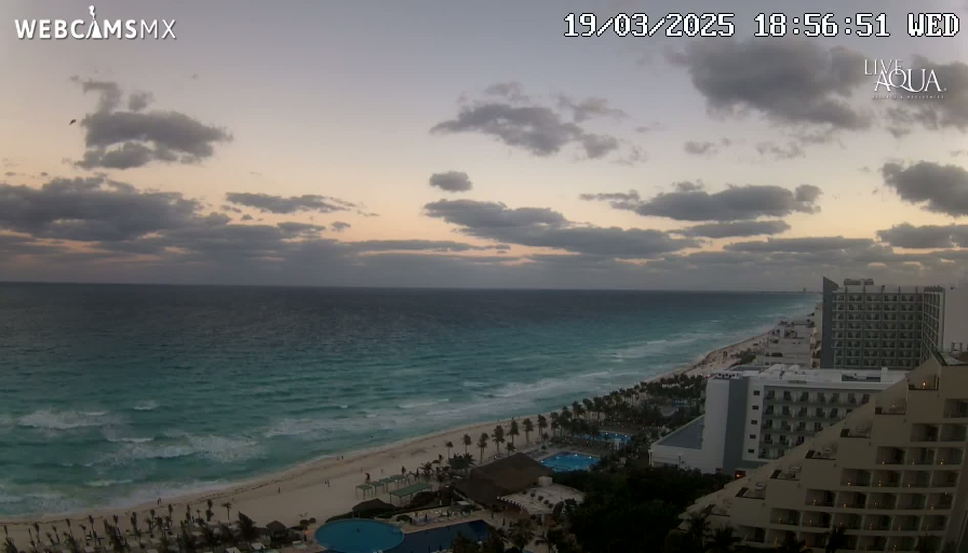 A partly cloudy sunset over a beach with turquoise water and several high-rise hotels.